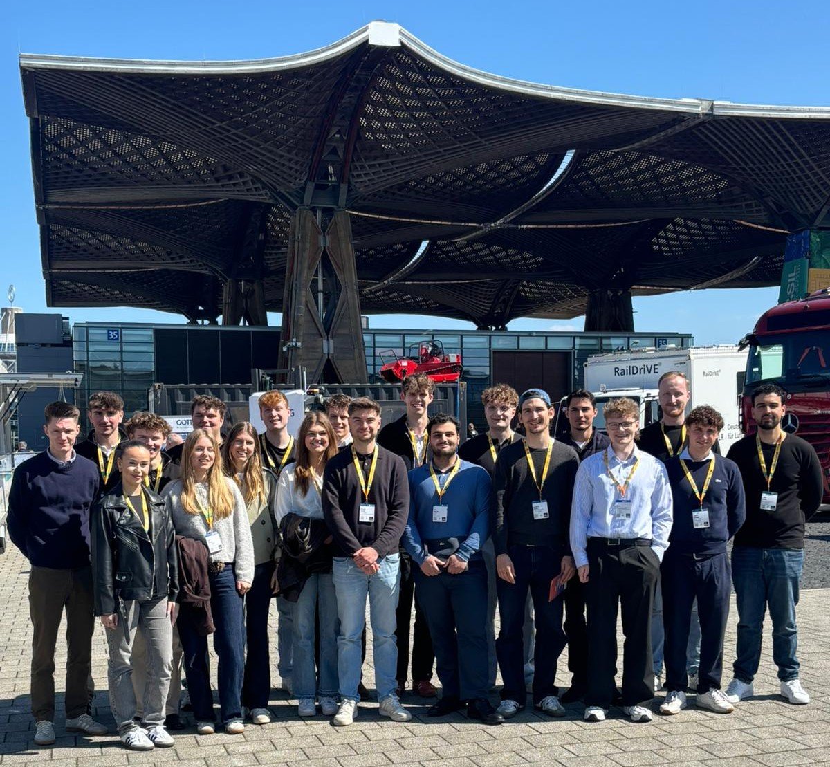 Eine große Gruppe von Studierenden der IBS Oldenburg posiert lächelnd während einer Exkursion vor einer markanten Holz-Dachkonstruktion auf einem Messegelände unter blauem Himmel.