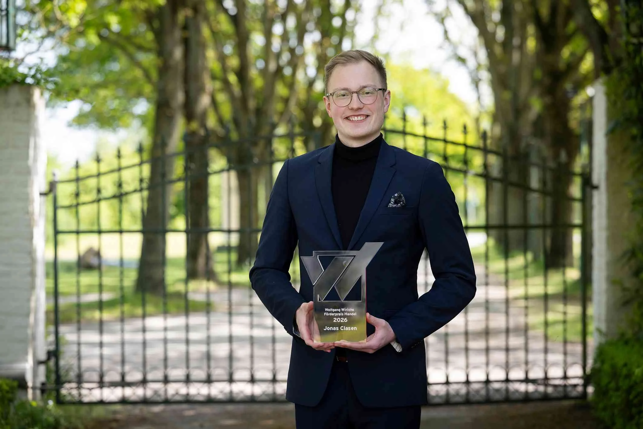 Jonas Clasen hält lächelnd den Wolfgang Wirichs Förderpreis Handel 2026 in den Händen. Er steht in einem dunklen Sakko vor einem schmiedeeisernen Tor im Grünen.