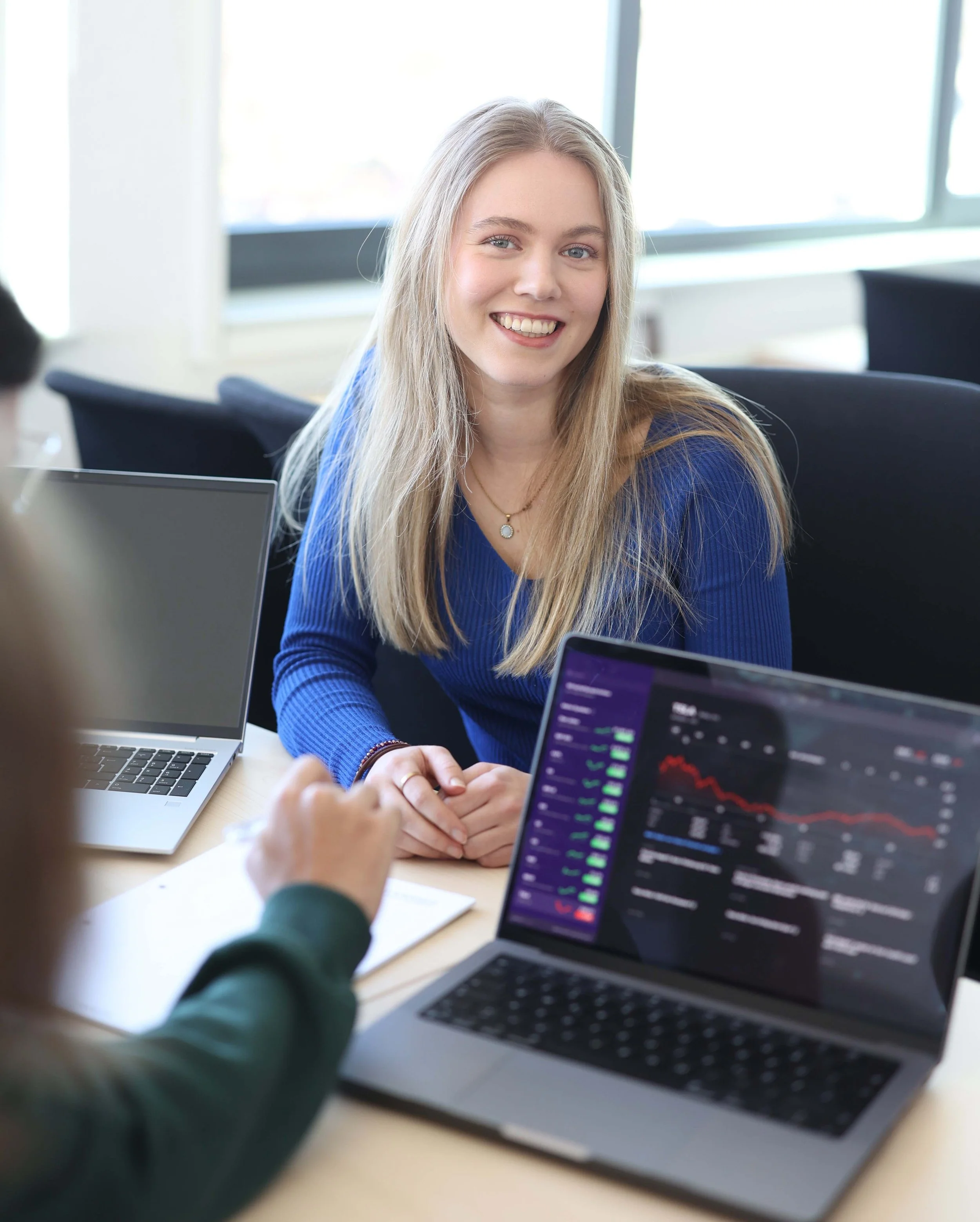 Eine lächelnde junge Frau im blauen Oberteil sitzt an einem Tisch bei der Gruppenarbeit. Vor ihr steht ein Laptop mit Datenvisualisierungen auf dem Bildschirm.