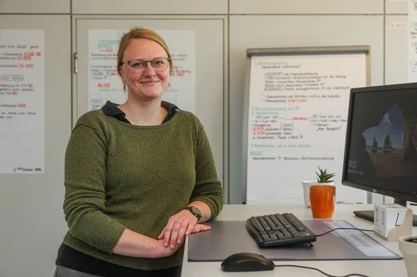 Eine lächelnde Frau mit Brille und grünem Pullover an einem Büroarbeitsplatz. Im Hintergrund hängen Flipcharts mit Notizen; auf dem Schreibtisch stehen ein Monitor und eine Tastatur.