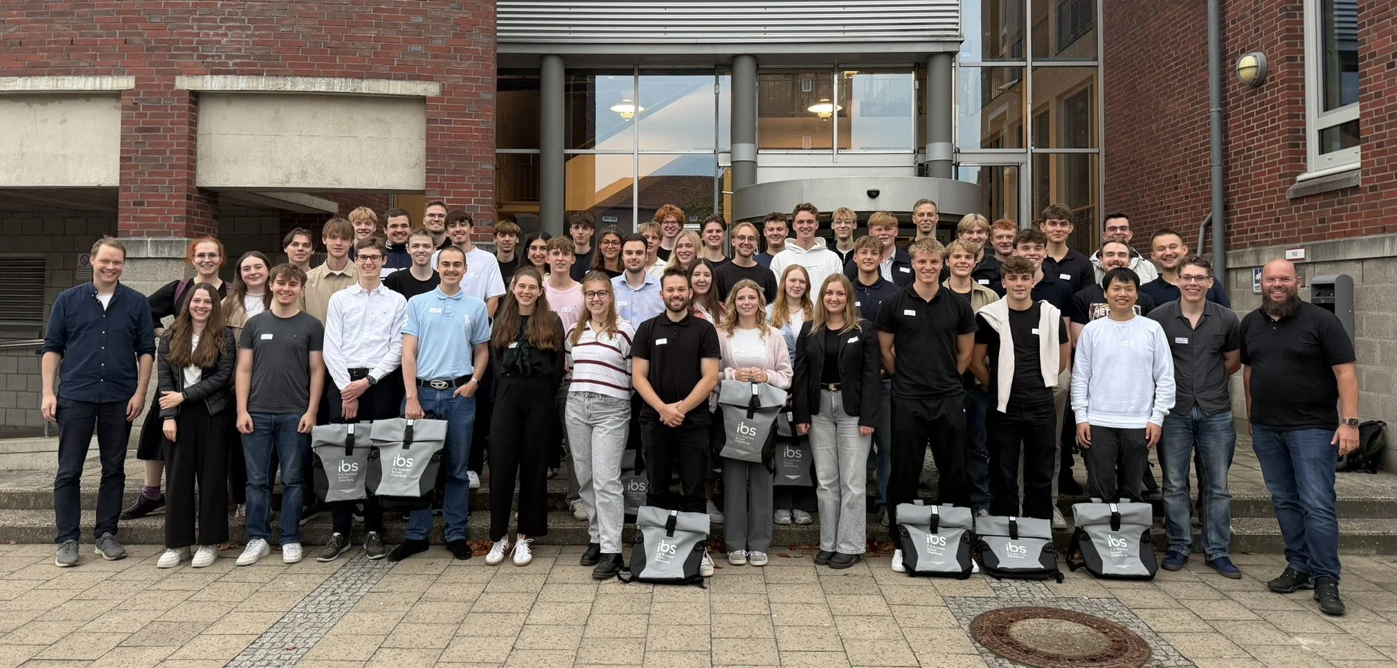 Eine große Gruppe neuer Studierender der IBS Oldenburg posiert für ein Gruppenfoto mit IBS-Rucksäcken vor dem Eingang des Hochschulgebäudes.