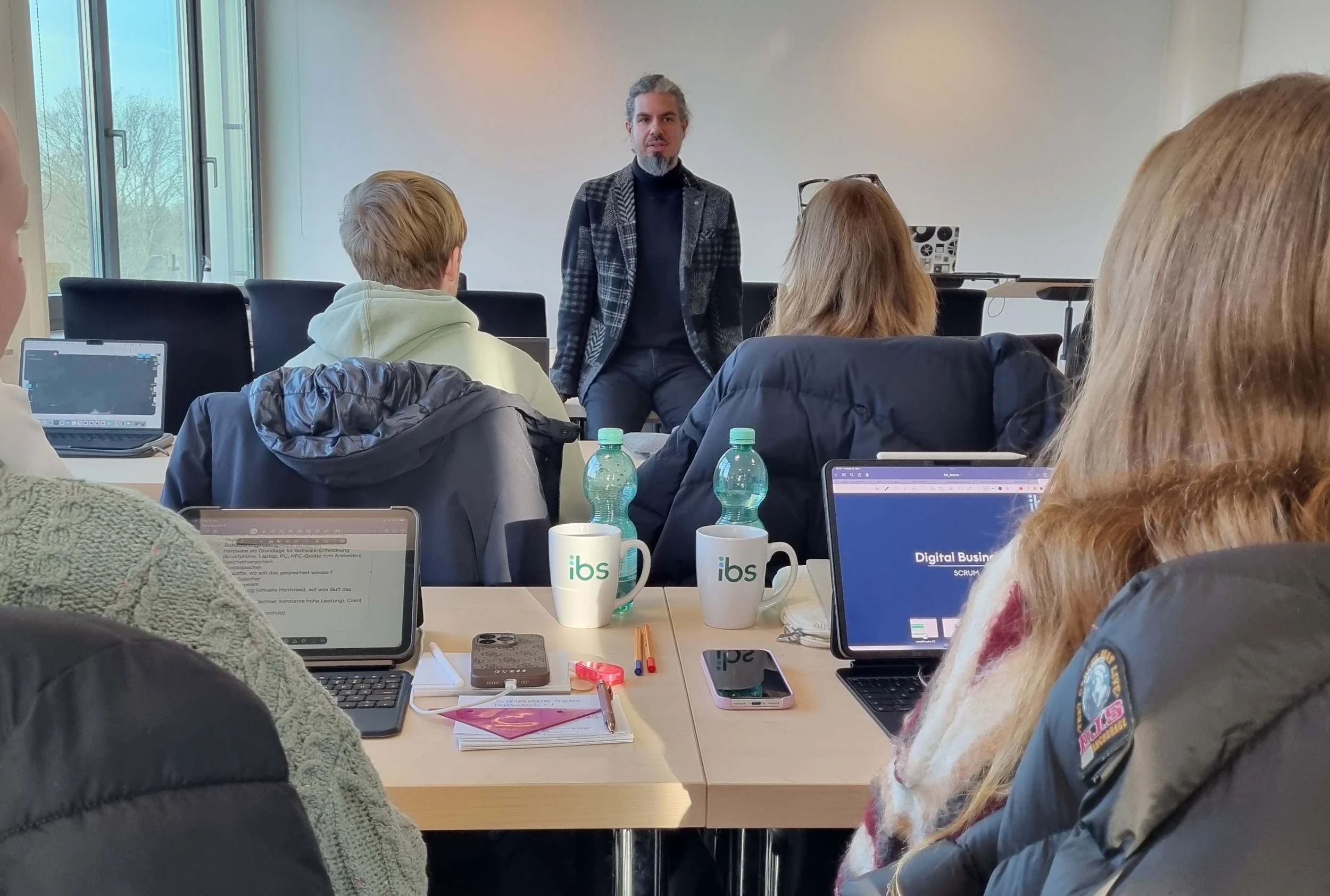 Dozent vor Studierenden in einer Vorlesung zu Digital Business an der IBS Oldenburg. Auf den Tischen stehen Tablets und Kaffeetassen mit IBS-Logo; die Atmosphäre ist modern und digital.
