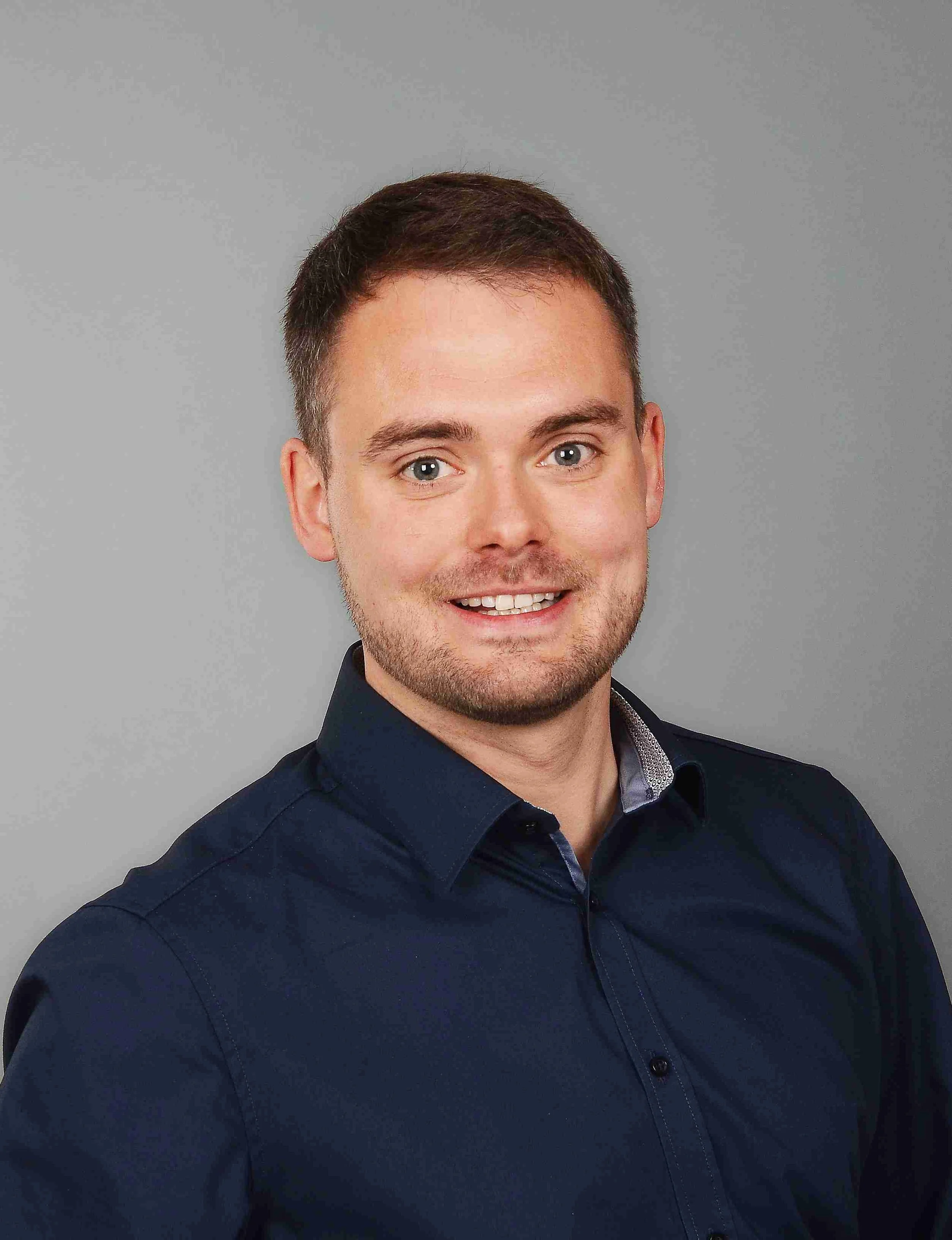 Professionelles Porträtfoto von Sebastian Lampe im dunkelblauen Hemd vor einem neutralen, grauen Hintergrund. Er blickt freundlich und lächelnd direkt in die Kamera.