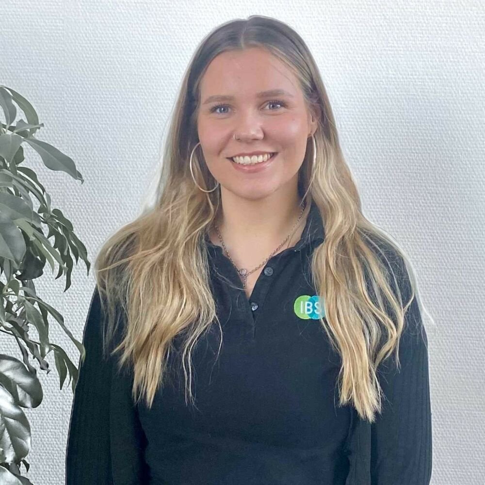 Anne Becker mit langem, lockigen Haar, großen Ohrringen und einem schwarzen Shirt mit einem IBS-Logo, stehend vor einer weißen Wand, neben einer grünen Pflanze, lächelnd.