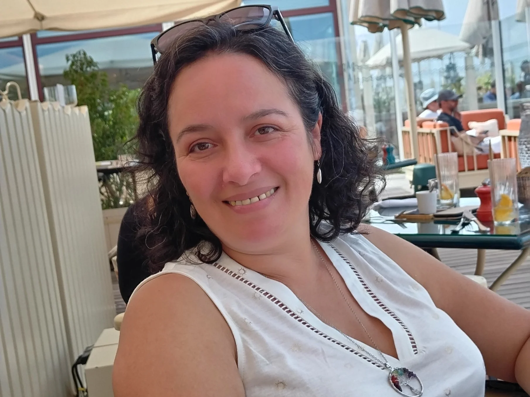 Une femme souriante avec des cheveux bouclés noirs porte un haut blanc et porte des lunettes sur la tête, assise dans un restaurant en plein air.