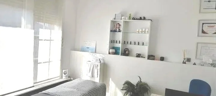 Une salle de soins ou de relaxation avec un lit, une étagère contenant des huiles essentielles ou des produits de soin, un tableau blanc, et une fenêtre laissant entrer la lumière naturelle.