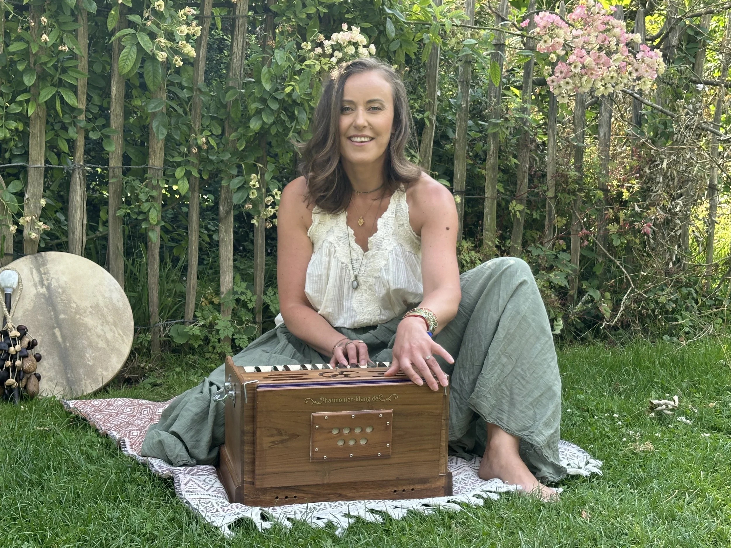 A woman with brown hair seated on the grass in a garden, playing a wooden harmonium. She is wearing a light-colored sleeveless top, light loose pants, and jewelry, and is smiling at the camera. Behind her, there is a wooden fence, pink and white flow