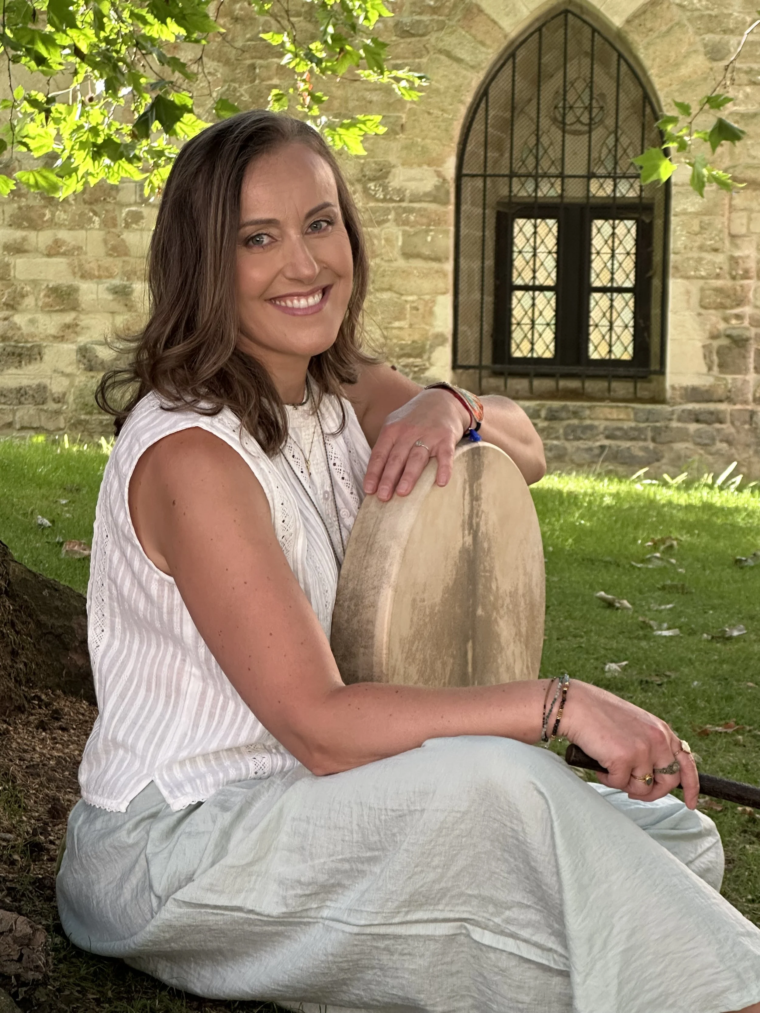 A smiling woman with shoulder-length brown hair sitting outdoors under a tree with green leaves. She is leaning on a large wooden object, possibly a musical instrument, with her arms resting on it. She is wearing a sleeveless white top, bracelets, an