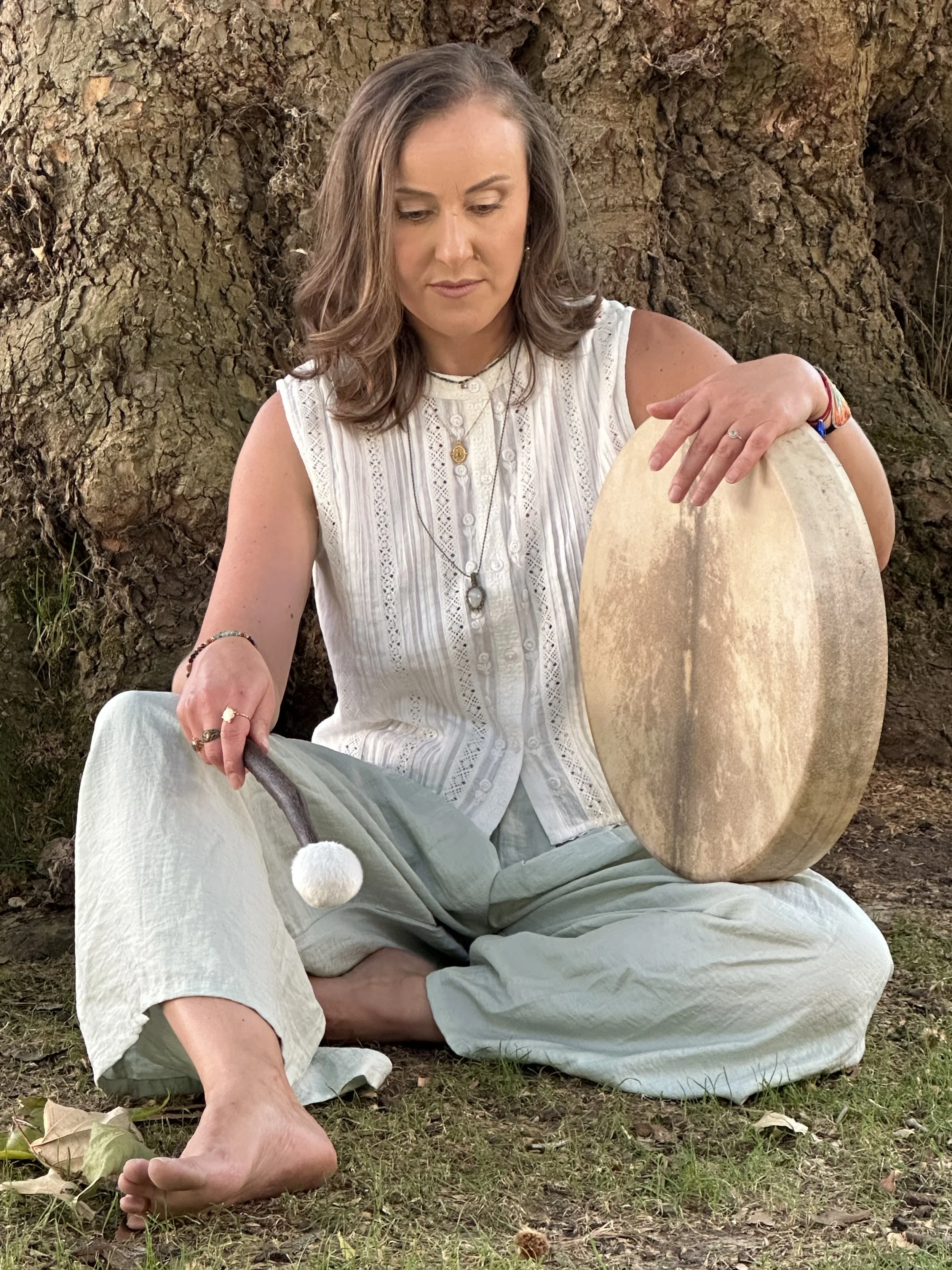 A woman sitting outdoors next to a large tree, holding a large drum, with her leg crossed underneath her and her foot resting on the grass.