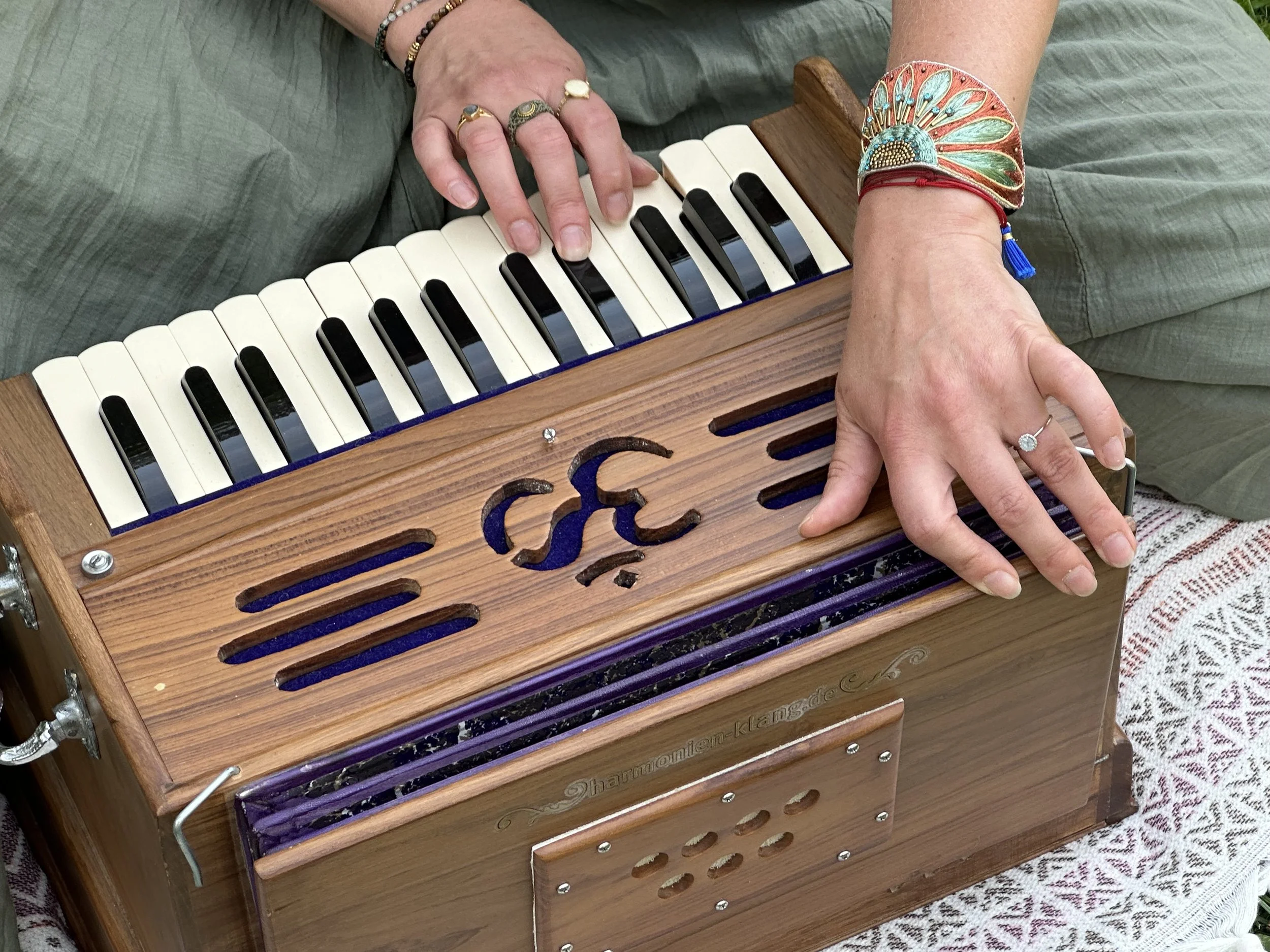 A person sitting on a green fabric surface playing a small wooden harmonica with their right hand, wearing multiple rings and colorful jewelry on their fingers and wrists.