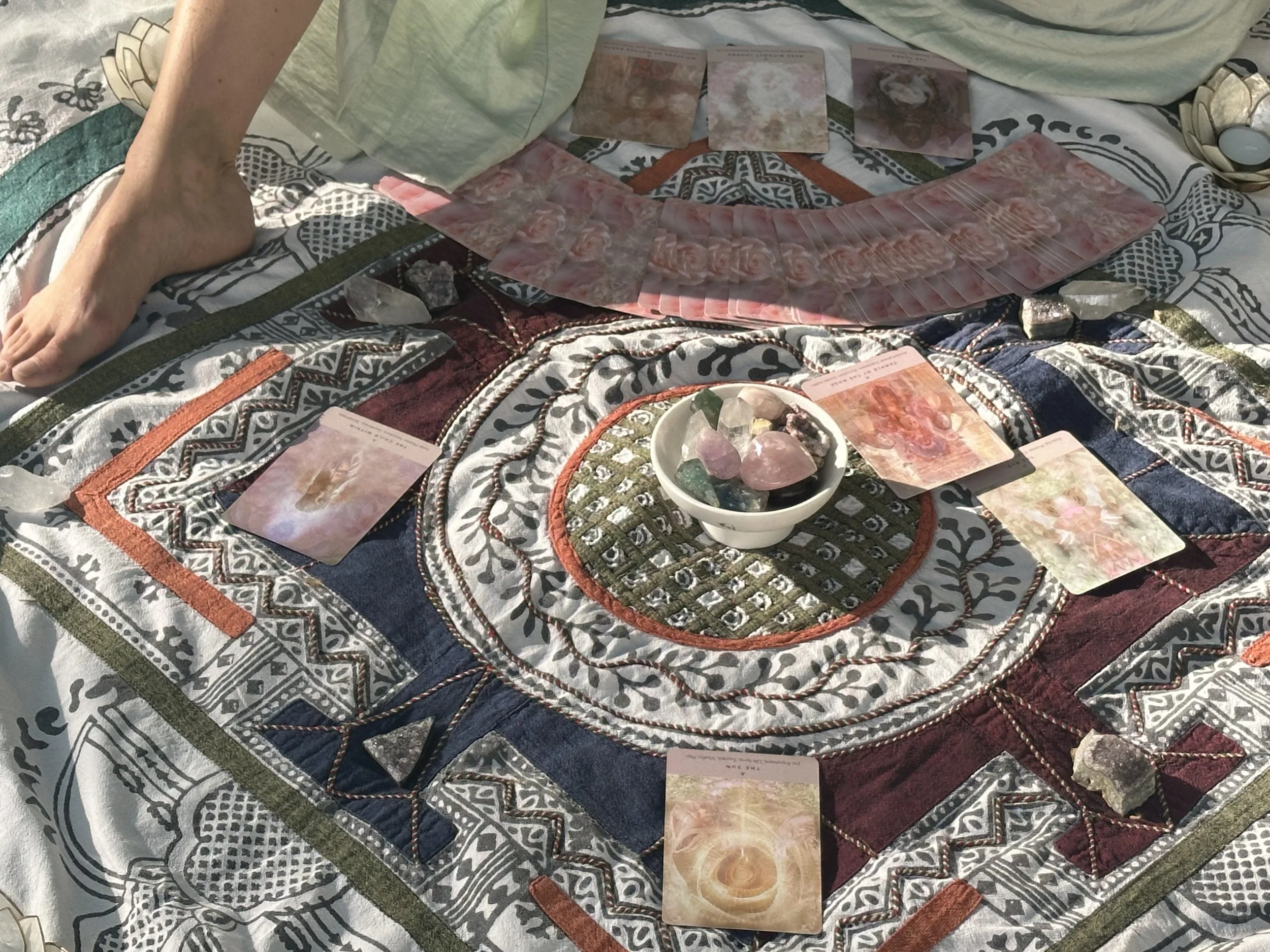 A person sitting on a patterned blanket with tarot cards laid out in a spread, including a fan of cards, and various crystals in a bowl, with other crystals surrounding the spread.