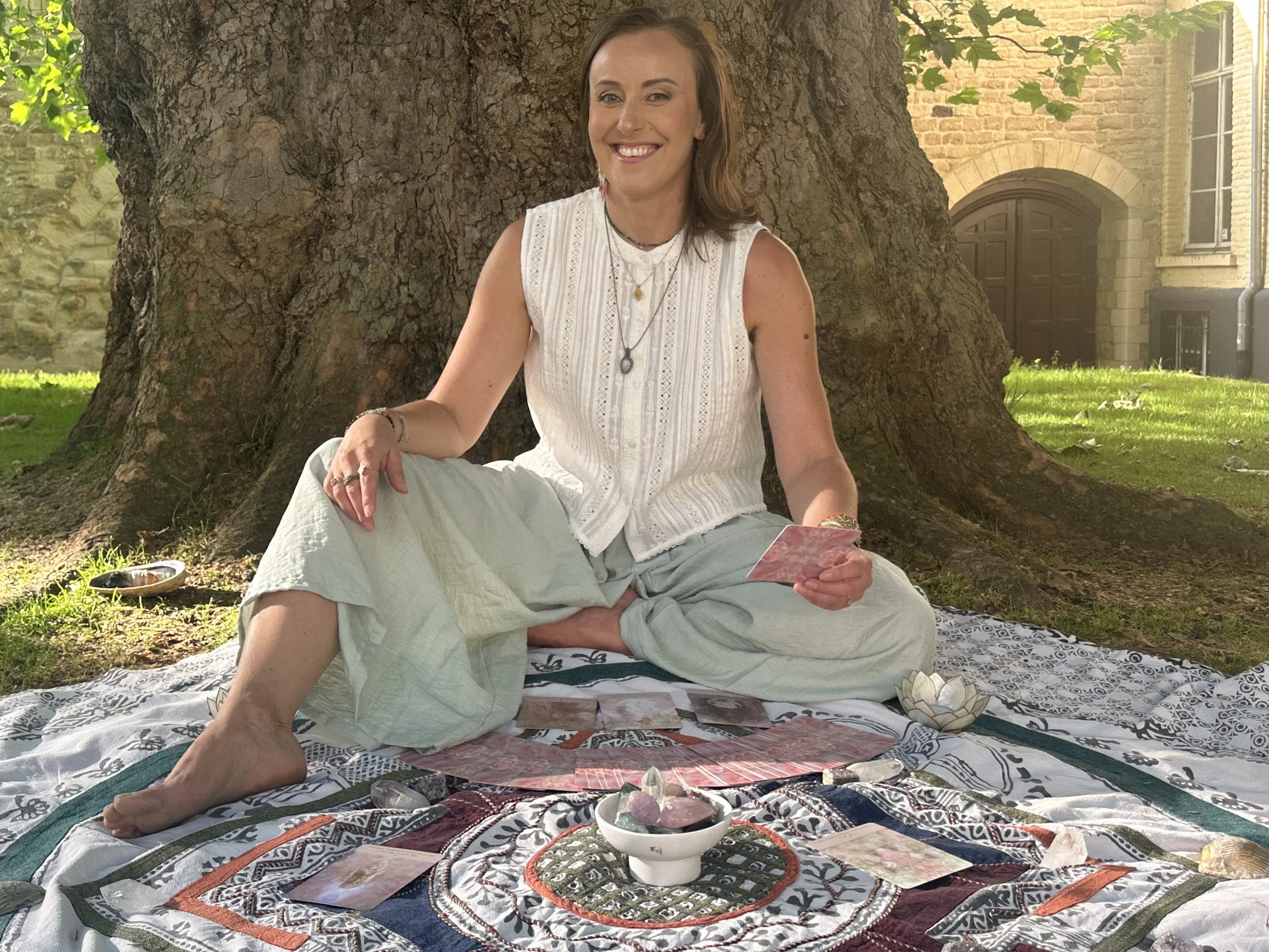 Woman smiling, sitting cross-legged on a patterned blanket beneath a large tree, holding tarot cards, with bowls of crystals and stones around her, in an outdoor setting near a brick building.