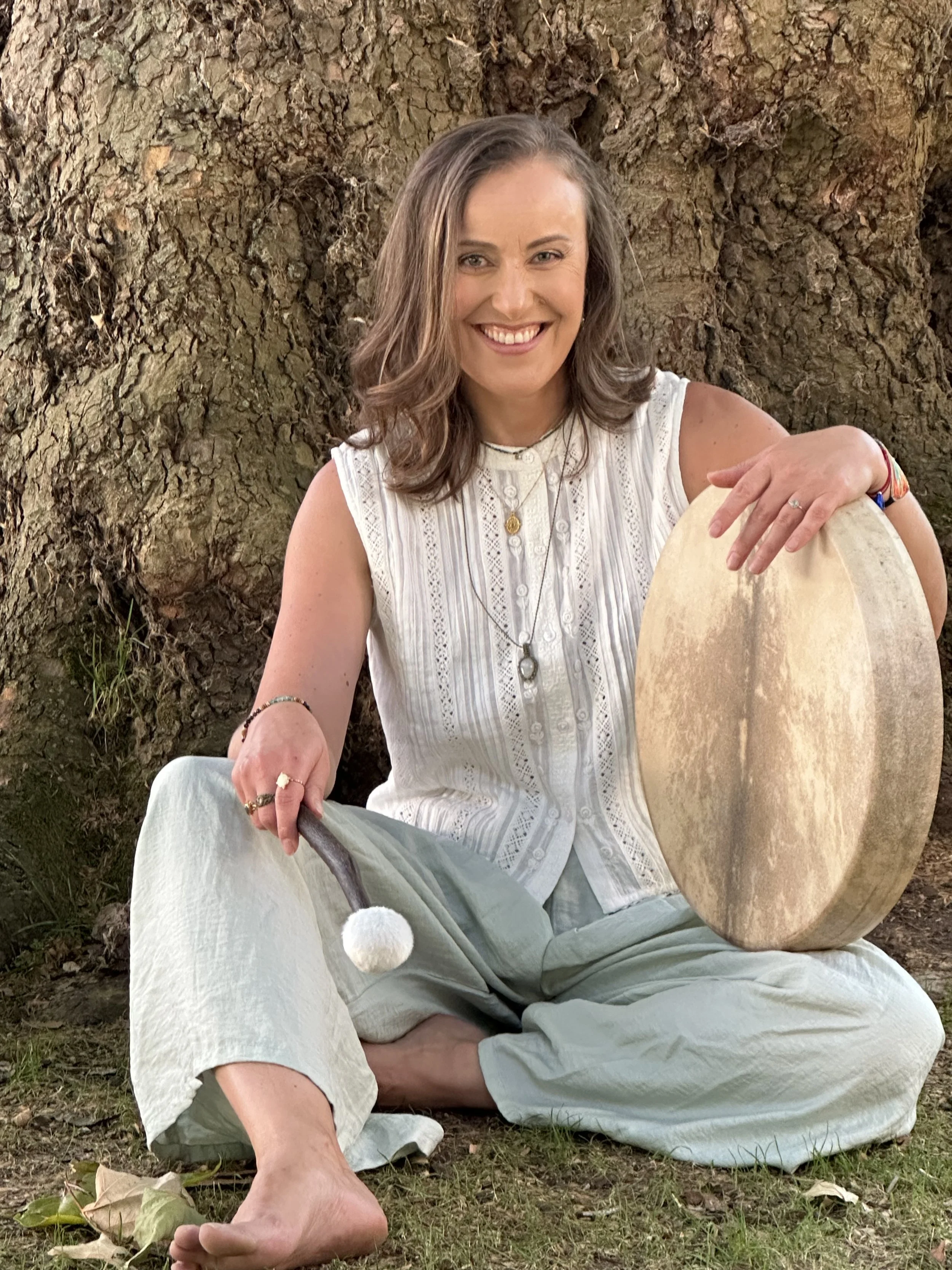 A woman sitting outdoors in front of a large tree with textured bark, smiling, wearing a white sleeveless top and light-colored pants, holding a large, round, flat object that looks like a drum, with her left hand resting on it.