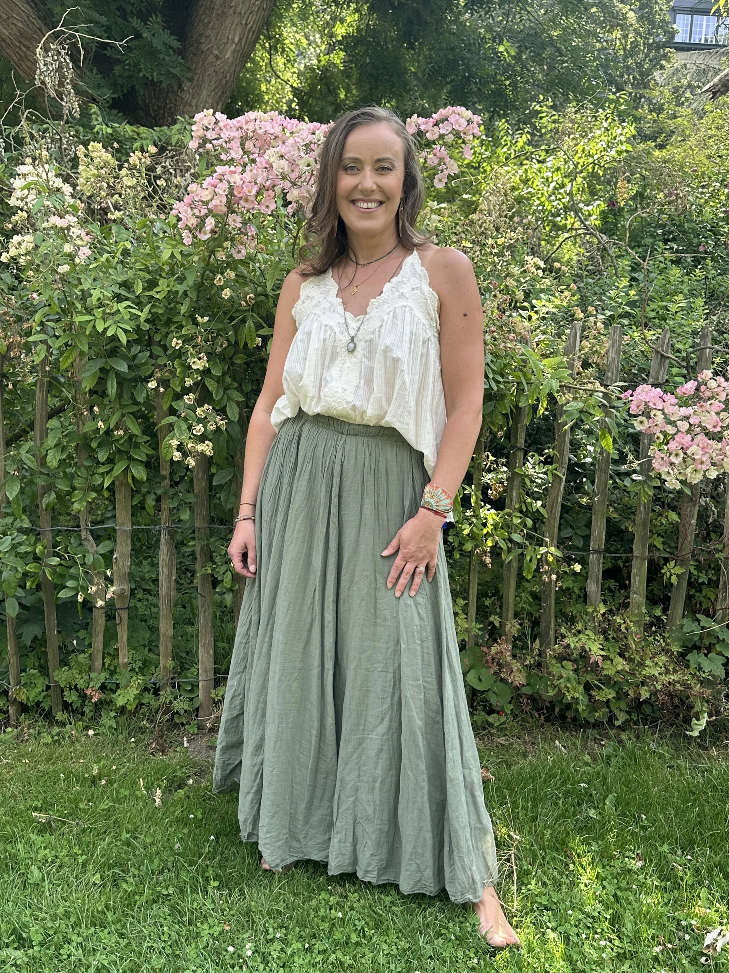 A smiling woman with brown wavy hair standing outdoors in front of a bush with pink flowers, a large tree, and a wooden fence. She is wearing a white sleeveless top, a long green skirt, and layered necklaces.