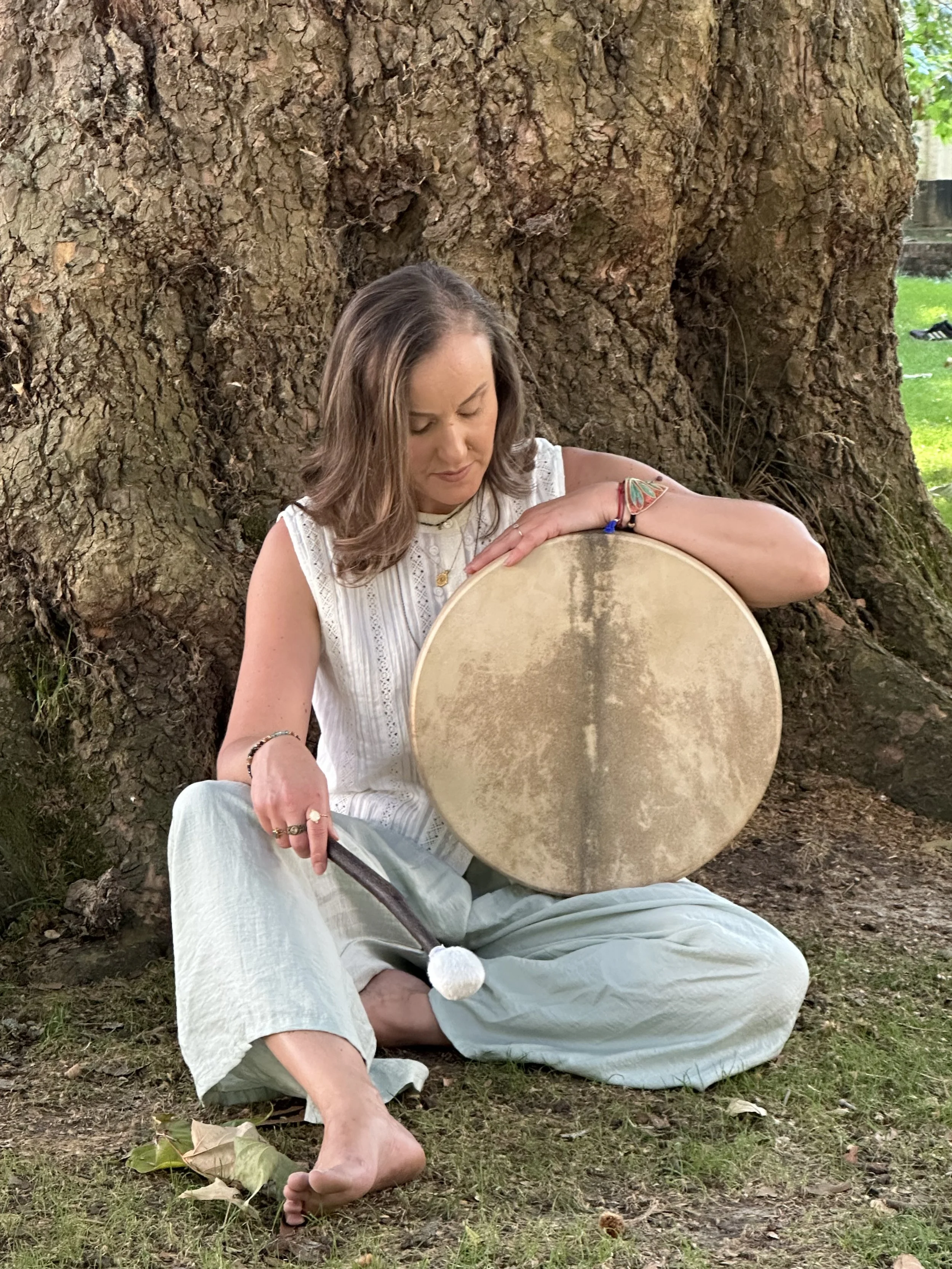 A woman with brown shoulder-length hair, wearing a white sleeveless top and light-colored pants, sitting on the ground outdoors in front of a large tree, holding a large round drum and looking down at it.