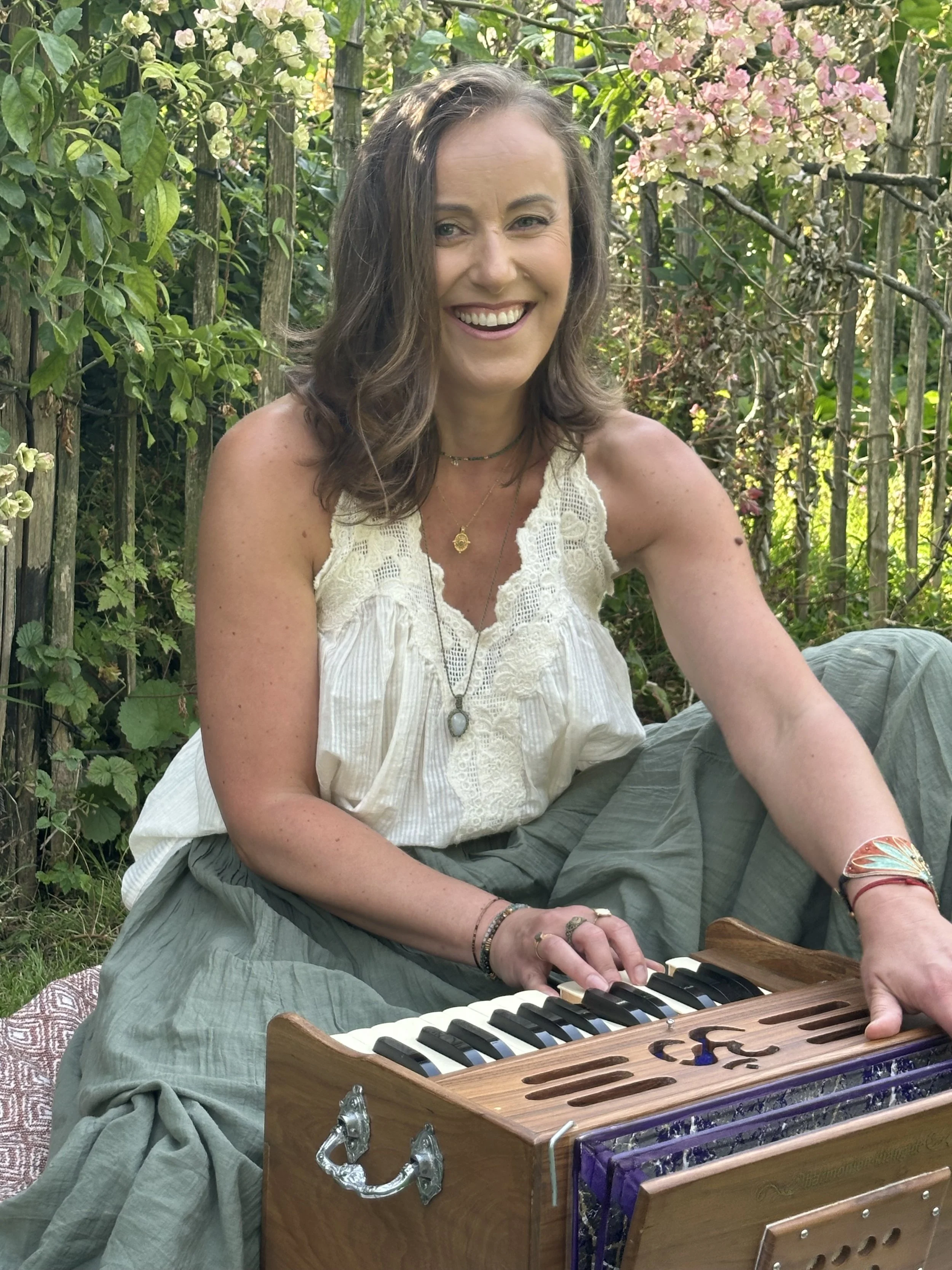 A woman with brown hair smiling and playing a wooden melodica outdoors, surrounded by greenery and pink flowers.