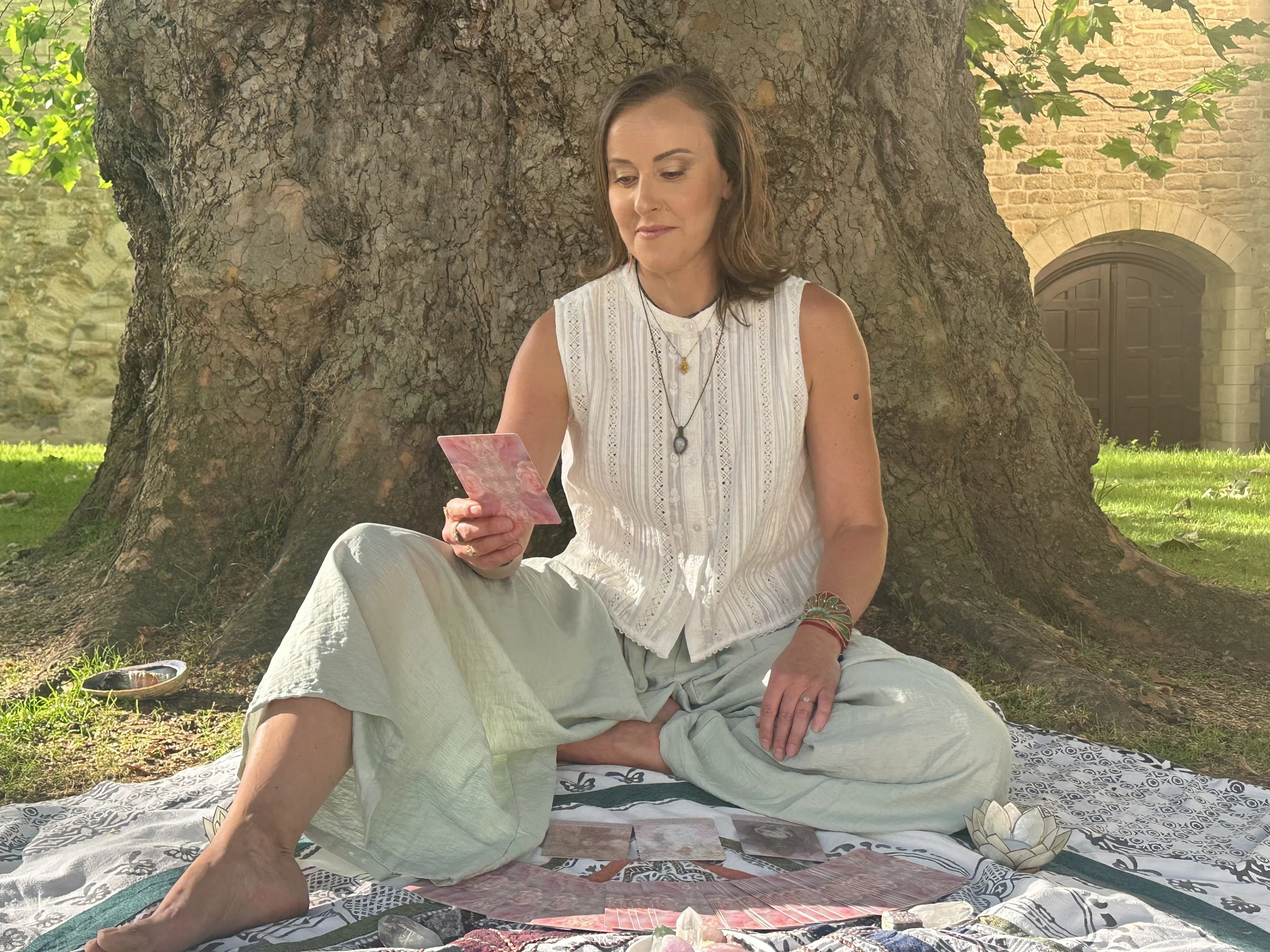 A woman sitting outdoors under a large tree, looking at a card in her hand, with tarot cards laid out in front of her on a blanket.