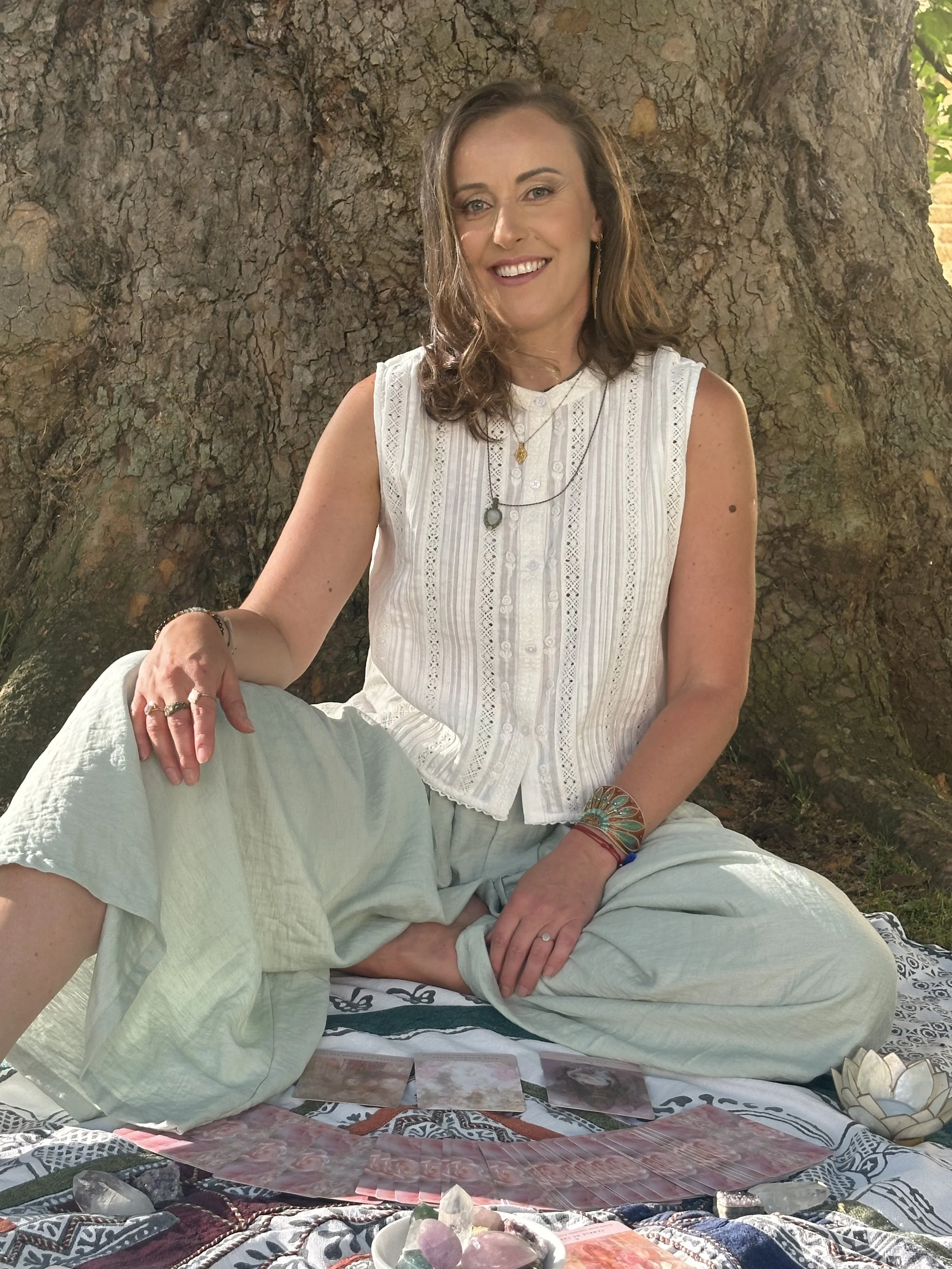 A woman with brown hair and earrings, wearing a white sleeveless blouse, is sitting outdoors in front of a large tree. She is smiling at the camera, with tarot cards and crystals laid out on a patterned cloth in front of her, indicating a tarot reading session.