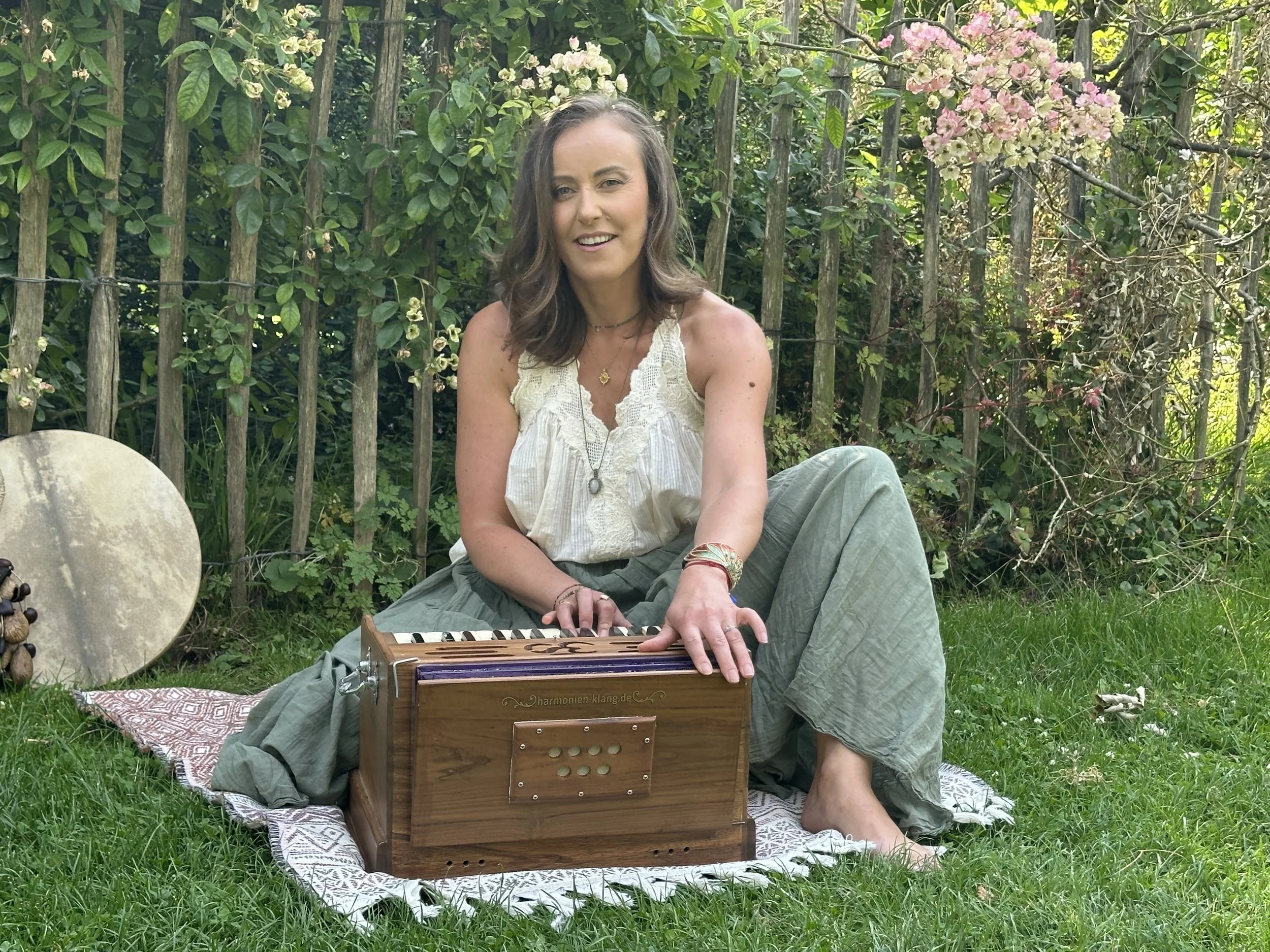 A woman with brown wavy hair in a white lace top sitting outdoors on a garden mat surrounded by greenery and flowers while playing a wooden harmonium with the brand name 'harmonien-klang.de' visible on it.