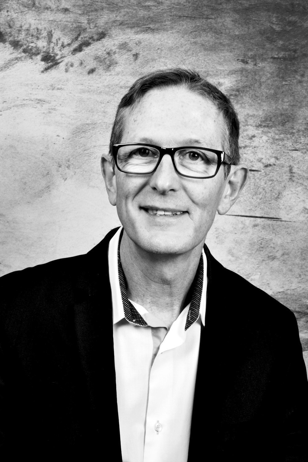 Portrait d'un homme portant des lunettes, souriant, avec des cheveux courts et une chemise bleue à pois clairs.