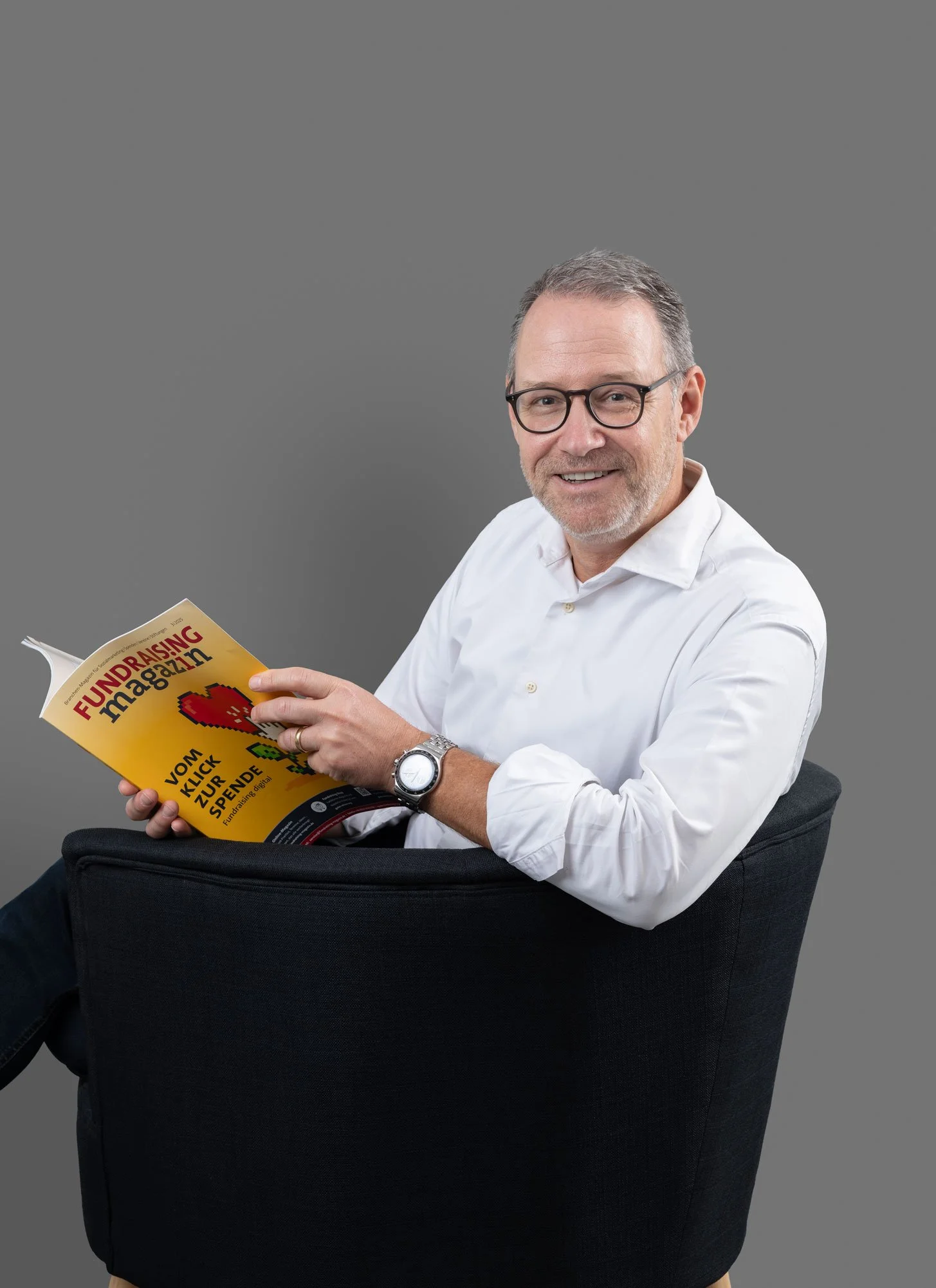 Ein Mann mit Brille, weißem Hemd und Armbanduhr sitzt in einem schwarzen Sessel, liest eine Zeitschrift mit dem Titel "Fundraising Magazin" gegen einen grauen Hintergrund.
