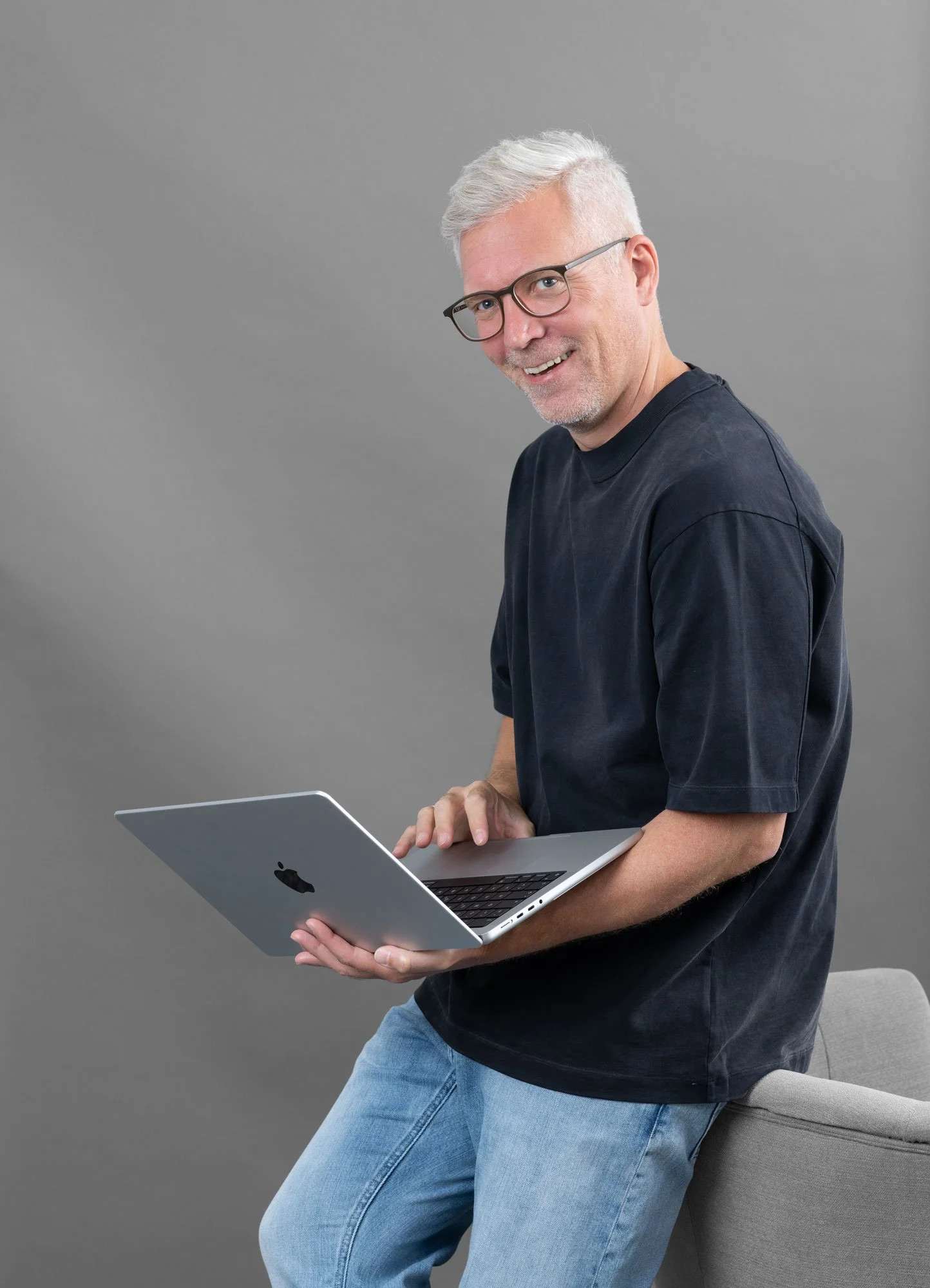 Ein älterer Mann mit Brille, grauem Haar und einem schwarzen T-Shirt hält einen offenen MacBook-Laptop in der Hand, steht vor grauem Hintergrund und lächelt.