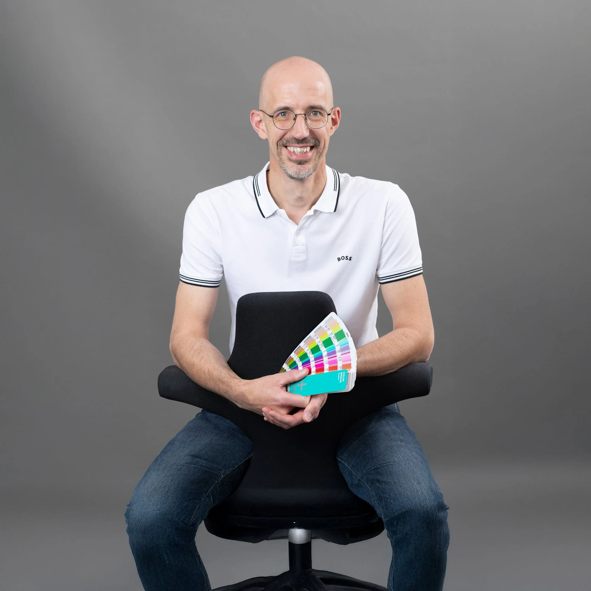 Ein lachender Mann mit Brille sitzt auf einem Stuhl vor einem grauen Hintergrund und hält Farbfächer in der Hand.
