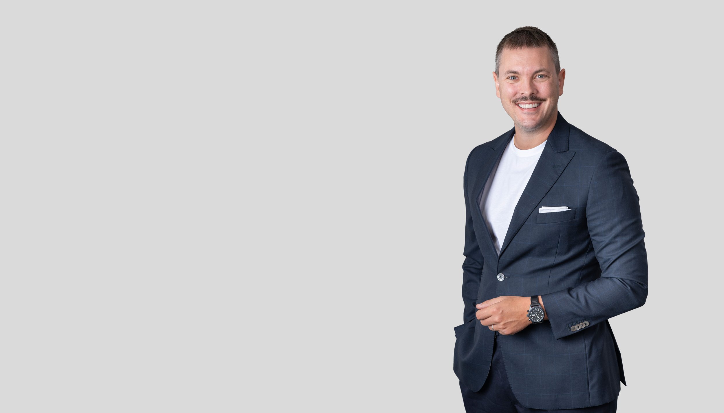Ein lächelnder Mann in einem dunklen Anzug mit weißem T-Shirt steht gegen einen hellgrauen Hintergrund