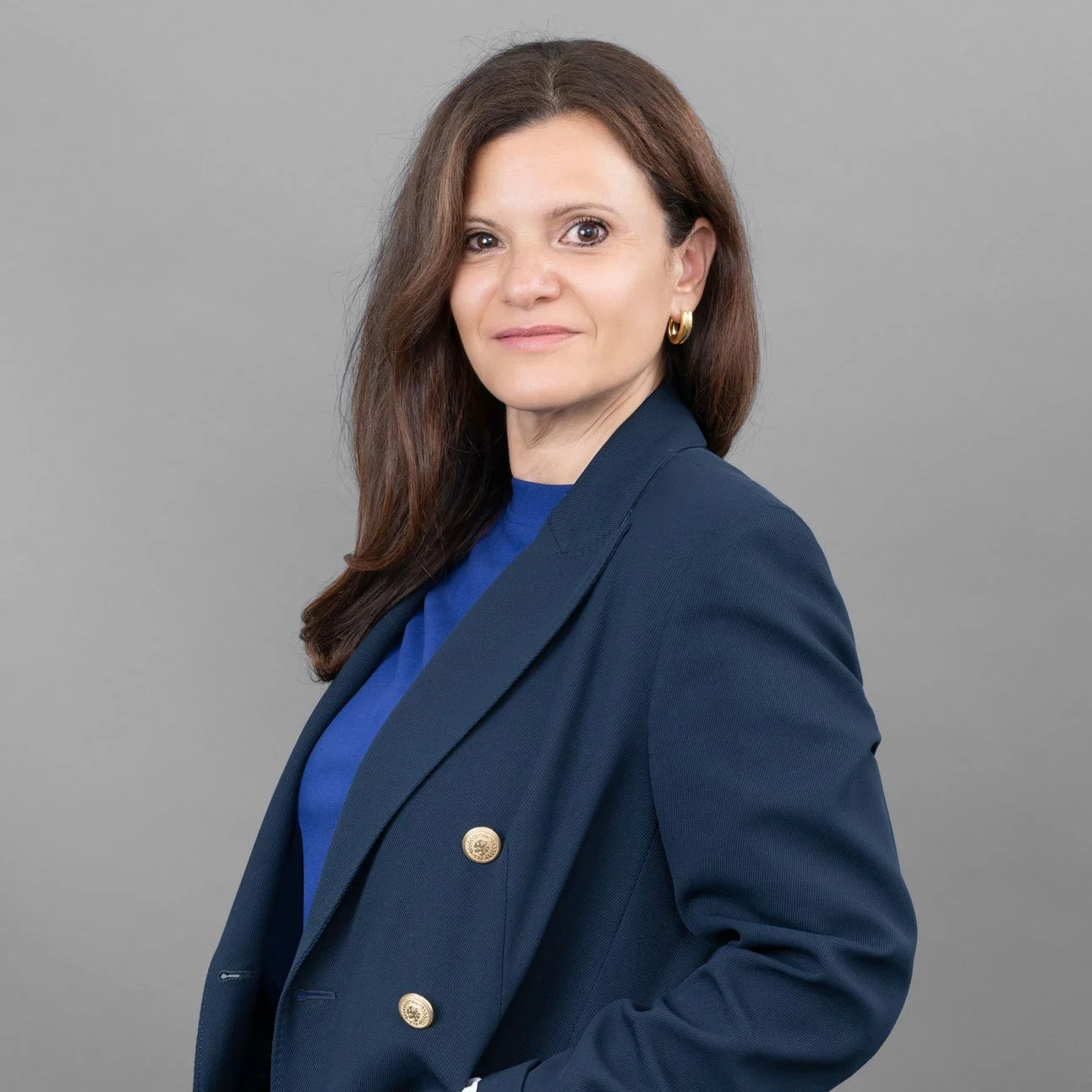 Porträt einer Frau mit braunen Haaren, blauen Kleidung und goldenen Ohrringen vor grauem Hintergrund.