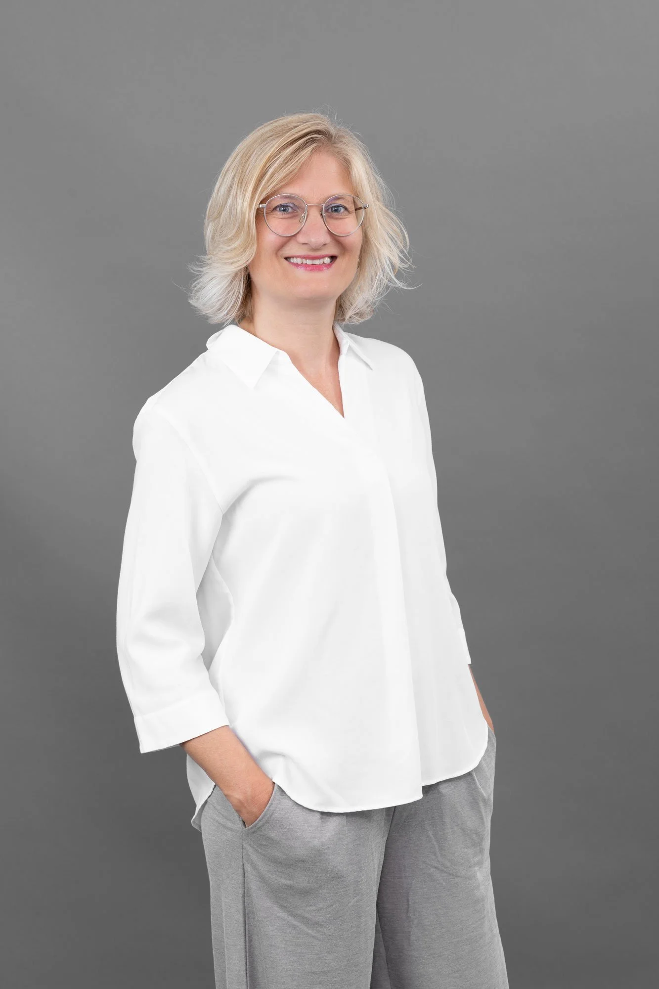 Eine lächelnde Frau mit blonden, lockigen Haaren, trägt eine weiße Bluse und graue Hosen vor einem grauen Hintergrund.