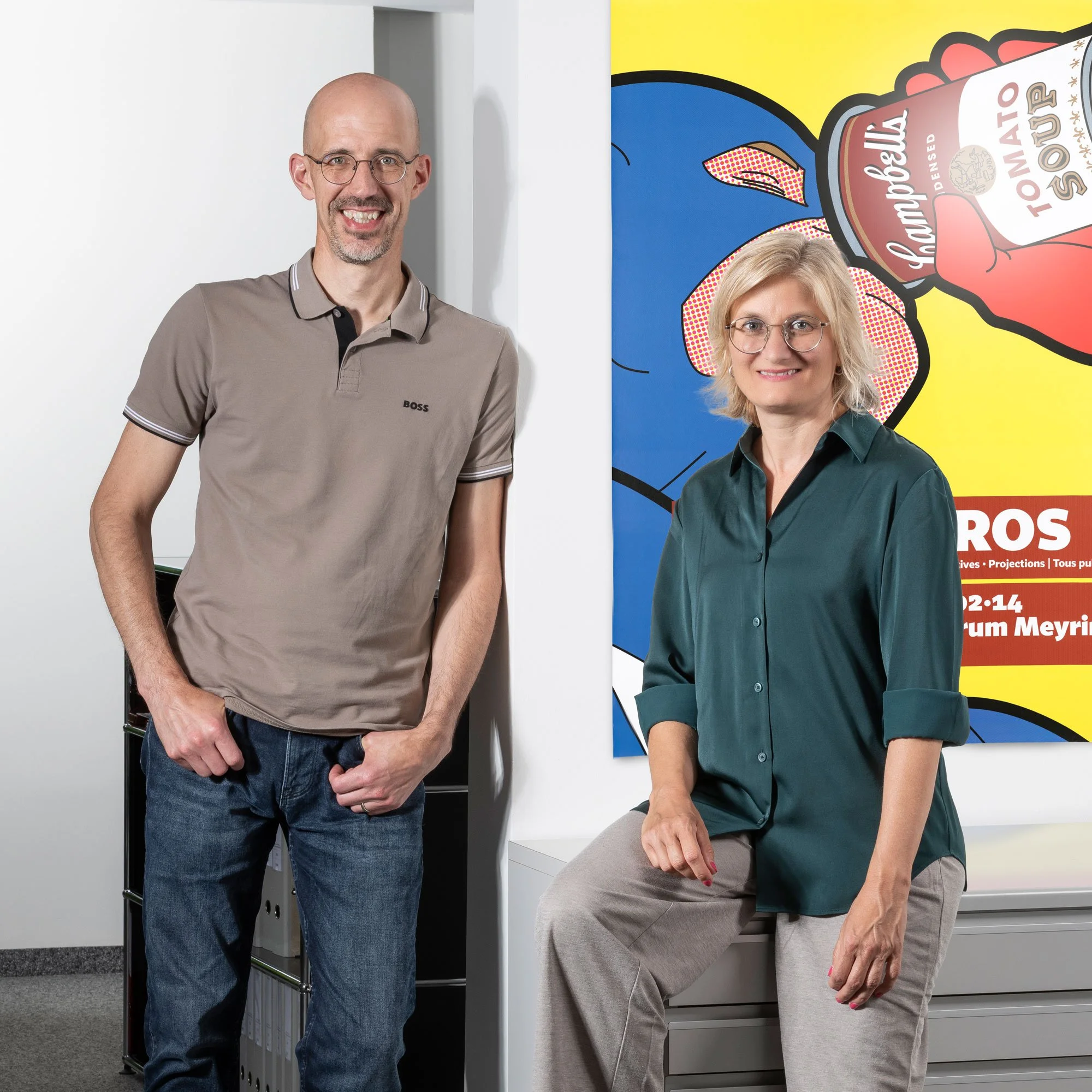 Ein Mann mit Brille, zwei Männer, und eine Frau, lächelnd vor einer bunten Wand mit einem Tomaten-Sugo-Plakat in einem modernen Büro.