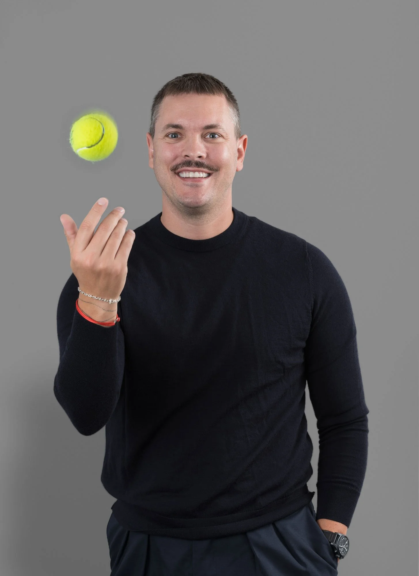 Ein lachender Mann mit kurzen Haaren und Schnäuzer, der einen Tennisball in der Hand hält, vor einem grauen Hintergrund.