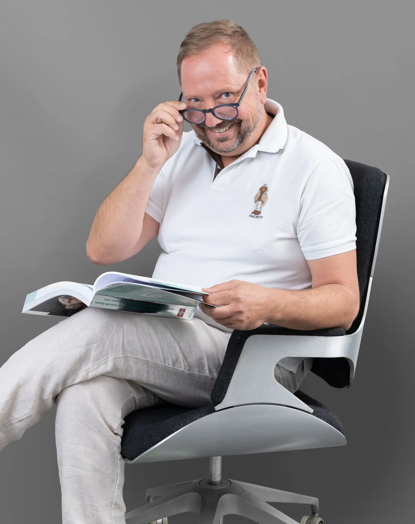 Ein Mann sitzt auf einem modernen Bürostuhl, trägt eine weiße Polo-Hemd und beige Hosen, liest eine Broschüre und trägt eine Brille, die er leicht anhebt.