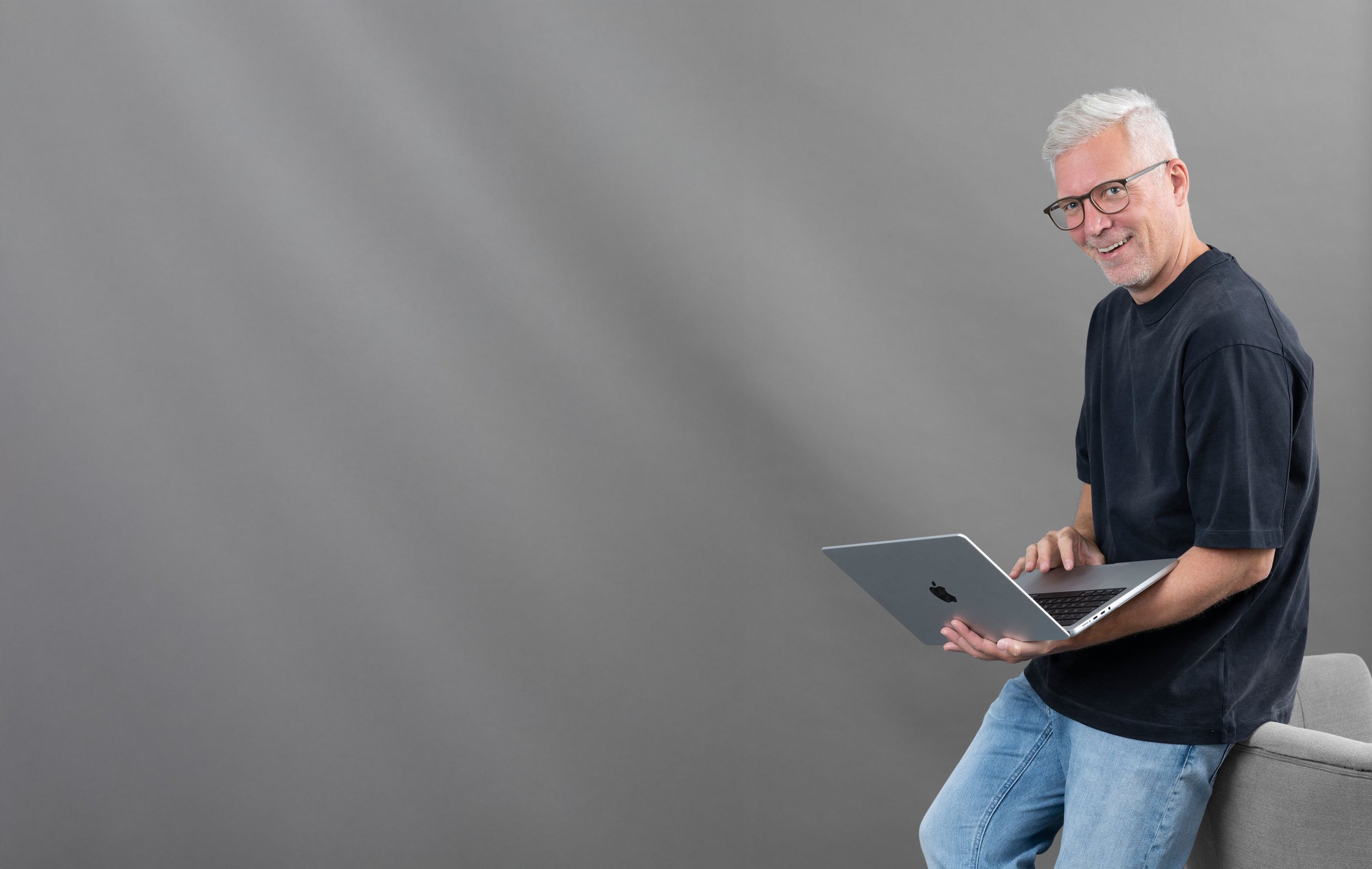 Ein älterer Mann mit weißen Haaren, Brille und dunklem T-Shirt, der auf einem grauen Stuhl sitzt und einen Laptop hält.