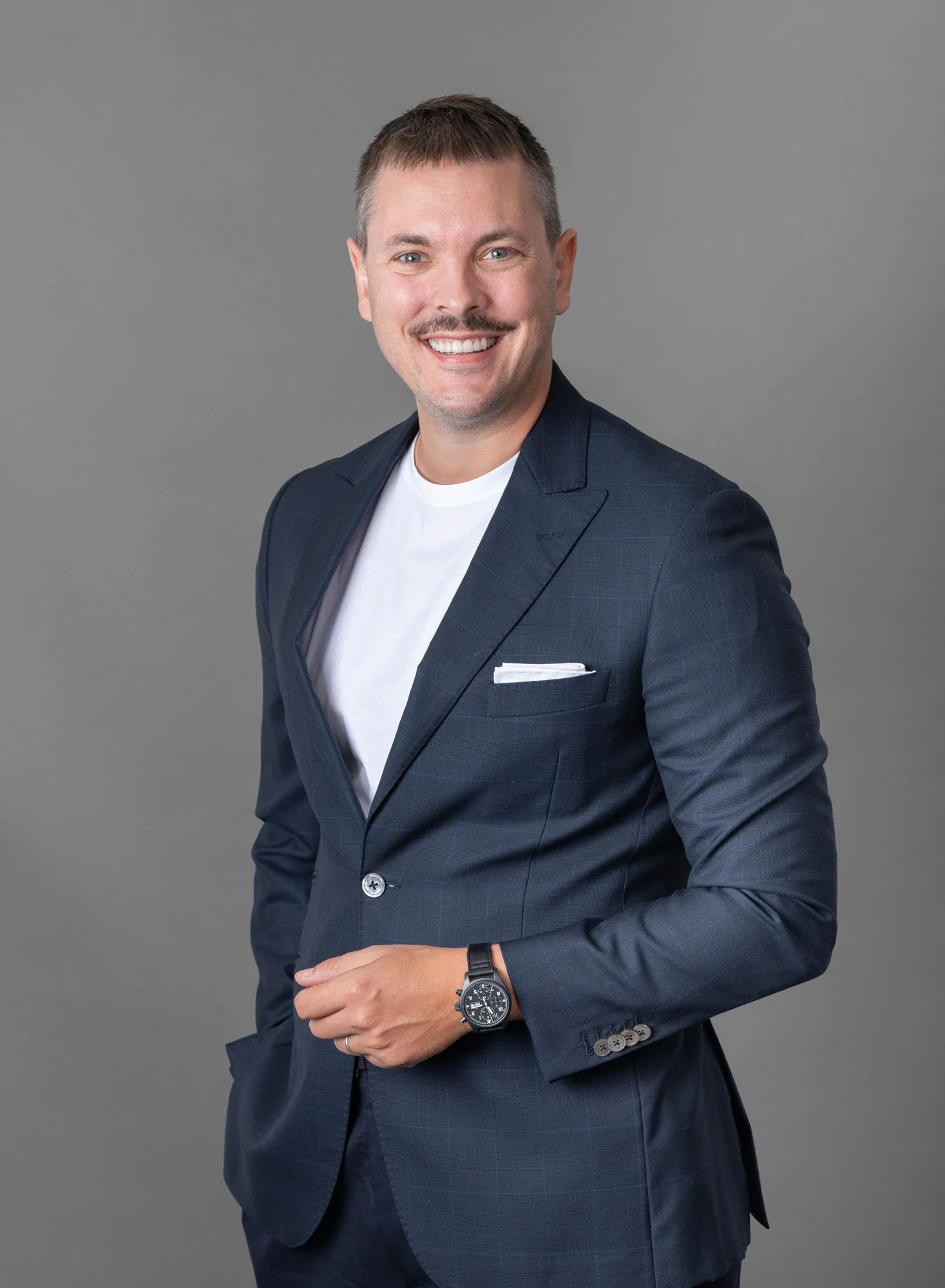 Ein lachender Mann in einem dunklen Anzug mit Krawatte, der vor einem grauen Hintergrund steht.