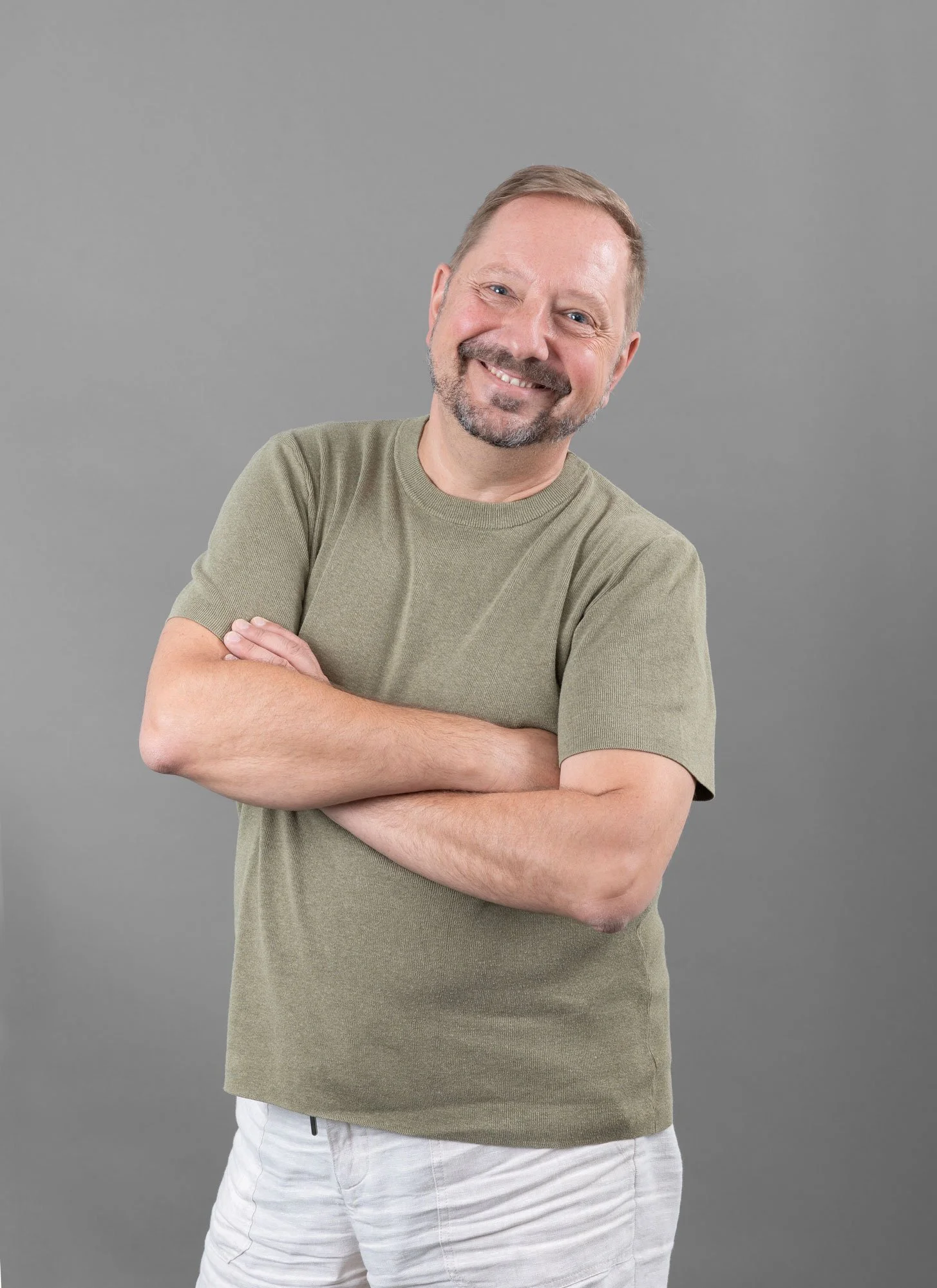 Ein lächelnder Mann mit kurzen braunen Haaren, Bart und Kinnbärtchen, trägt ein grünliches T-Shirt und weiße Hosen, steht vor einem grauen Hintergrund.