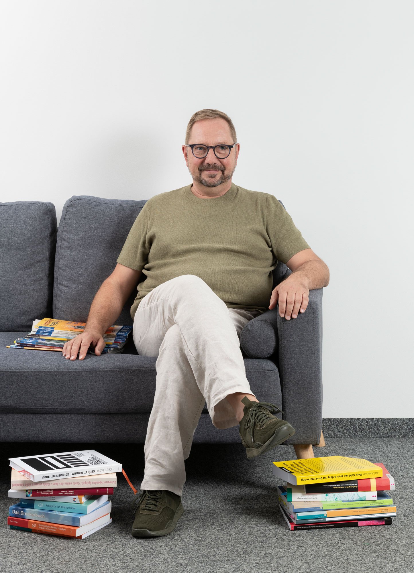 Ein Mann sitzt entspannt auf einem grauen Sofa in einem Raum mit weißer Wand, umgeben von aufgestapelten Büchern auf dem Teppichboden.