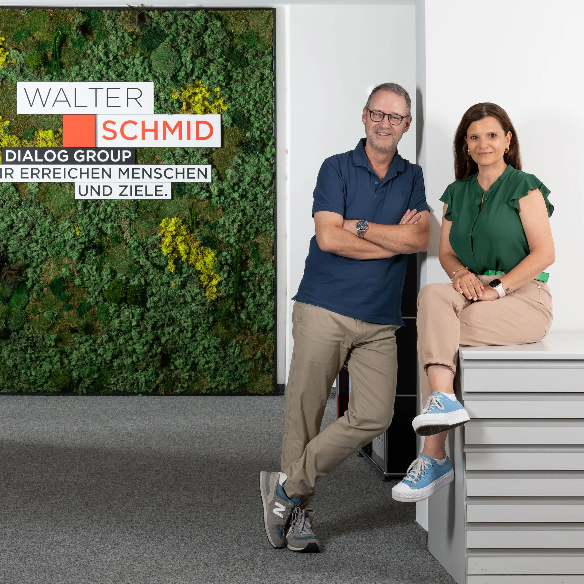 Zwei Personen stehen in einem modernen Büro neben einem großen Begrüßungsbild mit grüner Pflanzenwelt und Text. Der Mann trägt eine blaue Polo und beige Hosen, die Frau sitzt auf einer weißen Ablage, trägt ein grünes Hemd, beige Hosen und Sneakers.