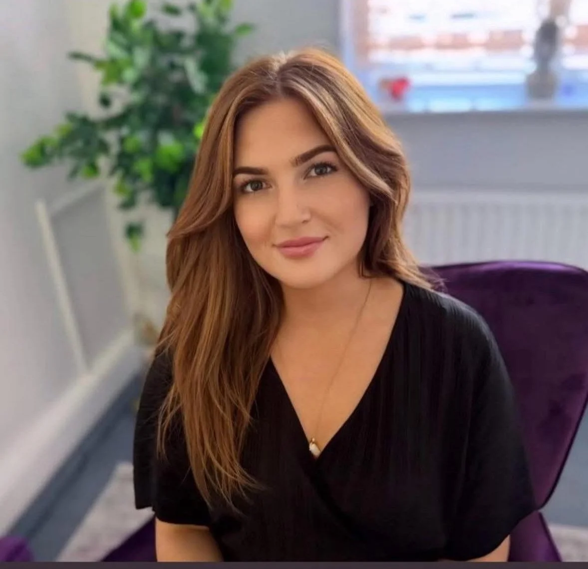 A woman with long, wavy brown hair and wearing a black top, sitting on a purple chair in a bright room with a potted plant and a window with sunlight in the background.
