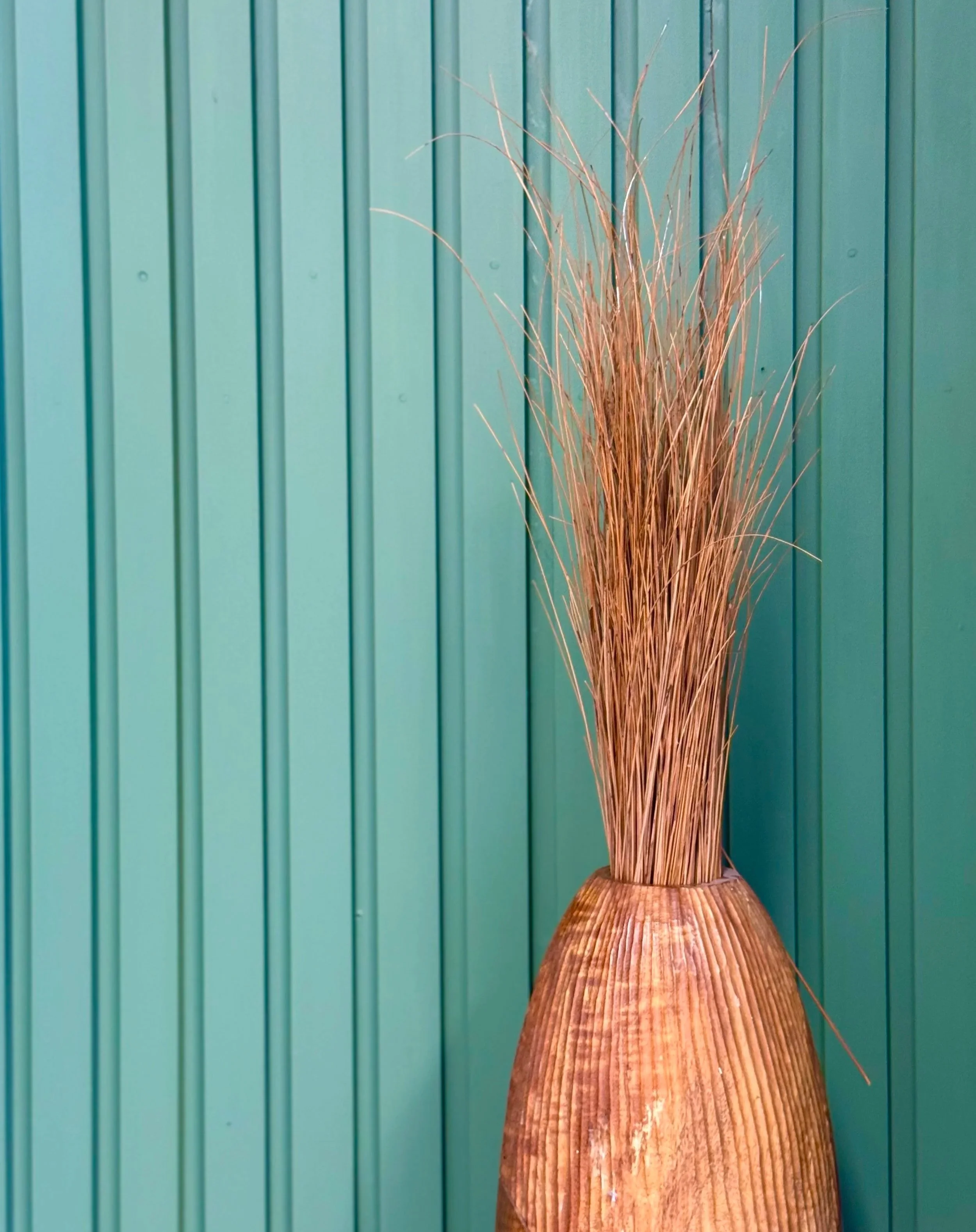 A tall wooden vase with a ridged surface holding dried ornamental grasses against a teal-colored vertical panel wall.