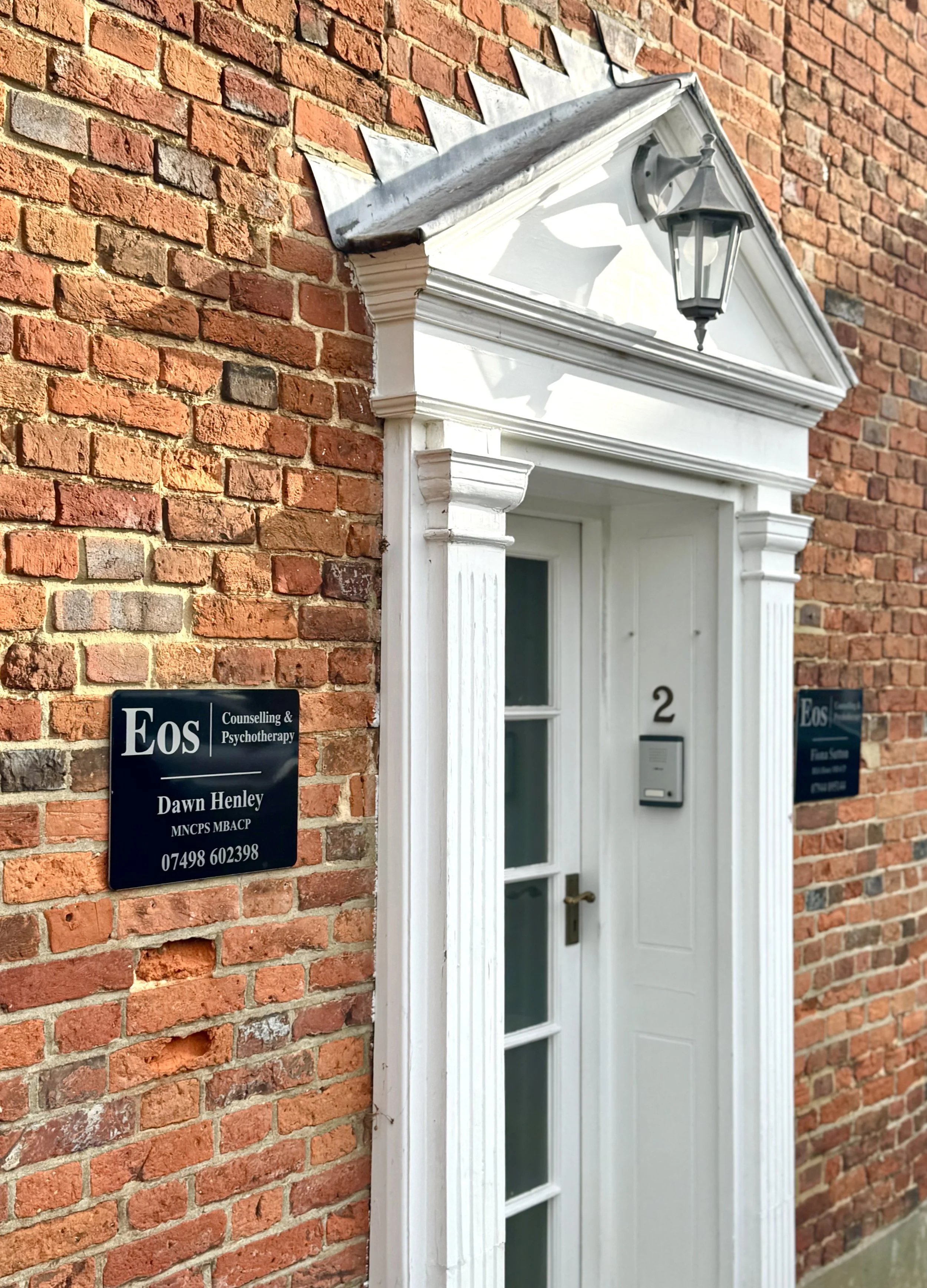 Brick building with white door, decorative trim, and lantern above the door, with signs for Eos Counseling & Psychotherapy on the wall.