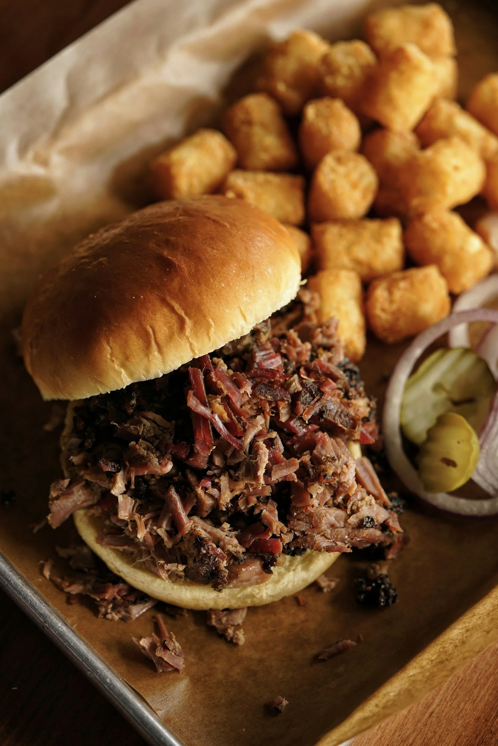 Barbecue brisket sandwich with toasted bun, topped with shredded barbecue beef, served with tater tots and pickles with onions on the side.