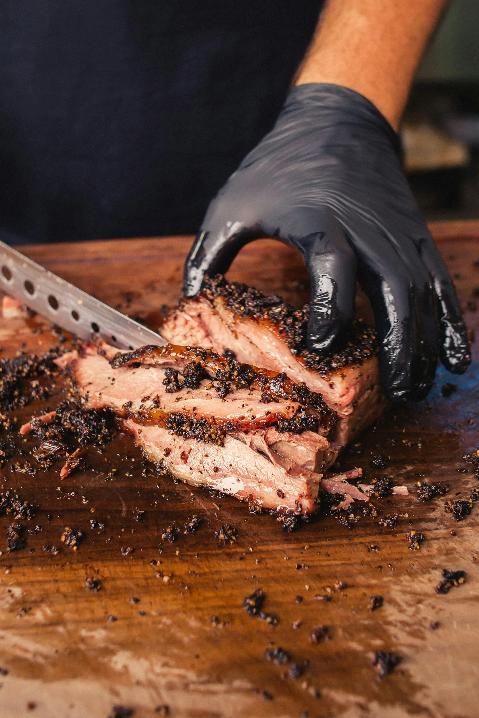 A person wearing black gloves slicing a smoked beef brisket on a wooden cutting board, with black pepper crust visible on the meat.