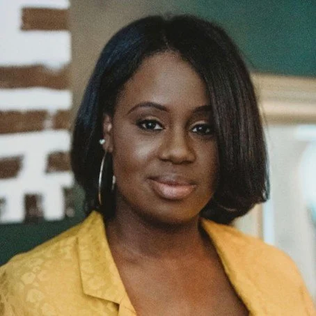 Portrait of a woman with shoulder-length black hair, wearing a yellow blazer, earrings, and makeup, standing indoors.