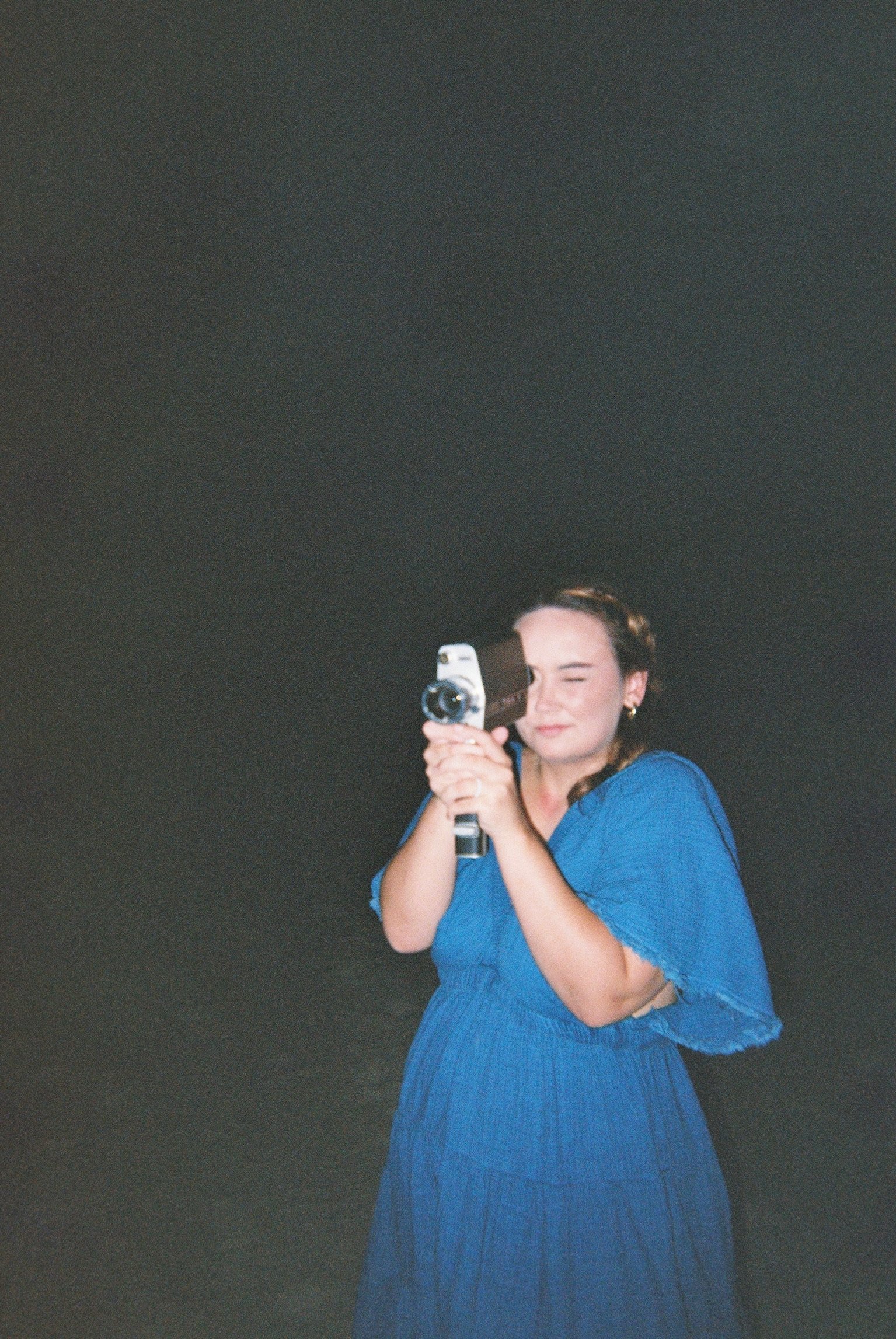 A woman with brown hair in a side braid, wearing a blue dress, holding a camera and pointing it towards the mirror. The background is dark and the woman appears to be taking a selfie.