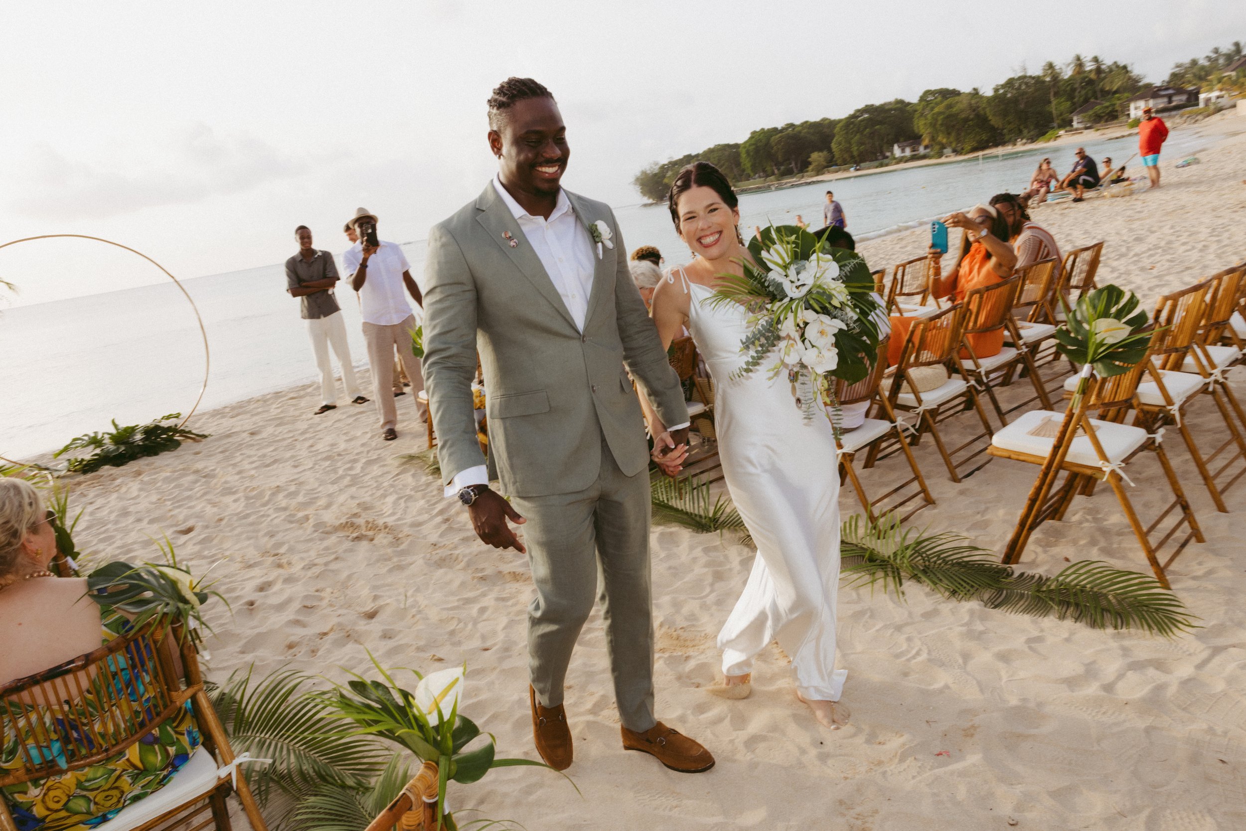A happy couple walks hand-in-hand on a sandy beach during their wedding ceremony, surrounded by seated guests and scenic water views.