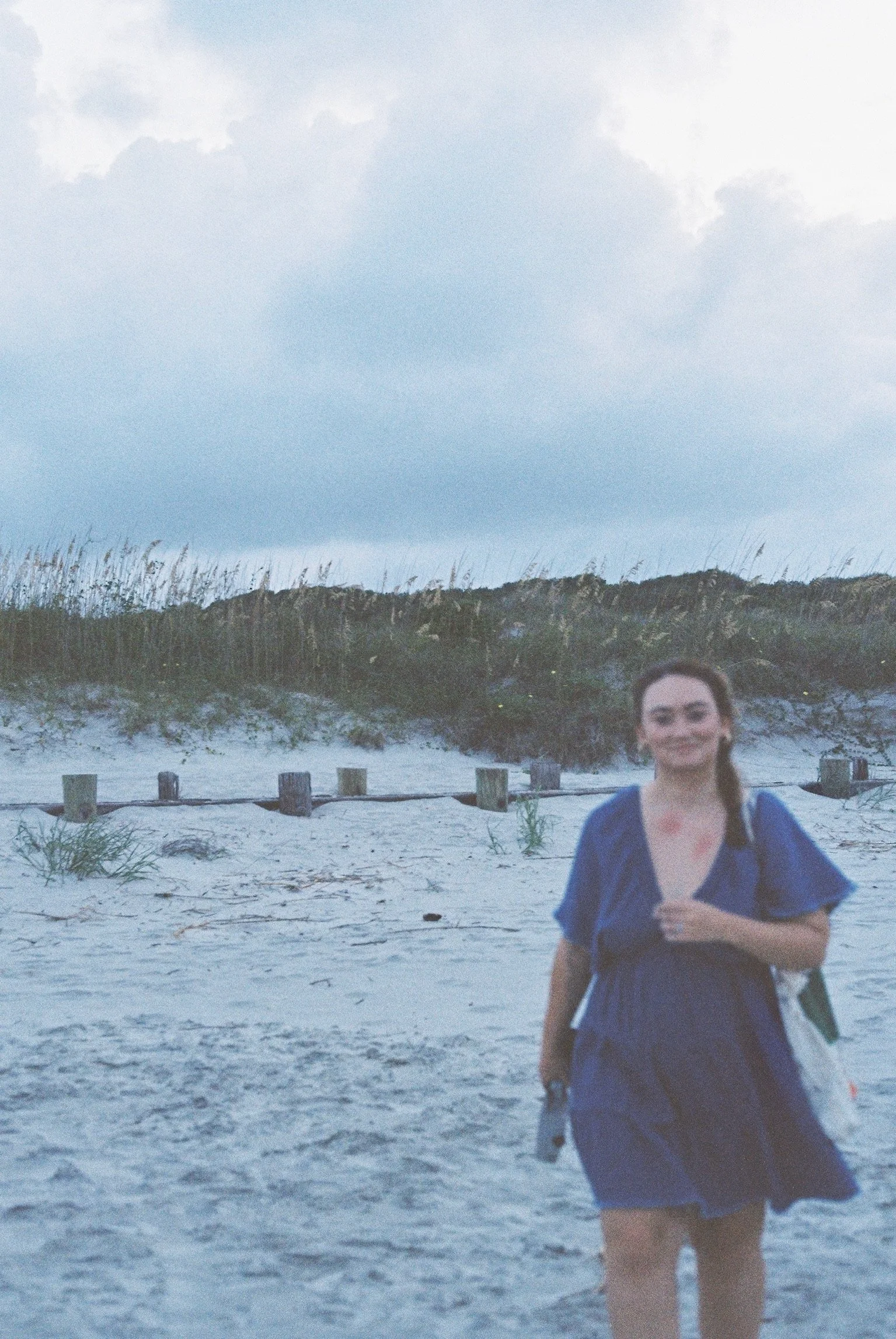 A woman smiling and walking on a sandy beach with dunes and sky in the background.