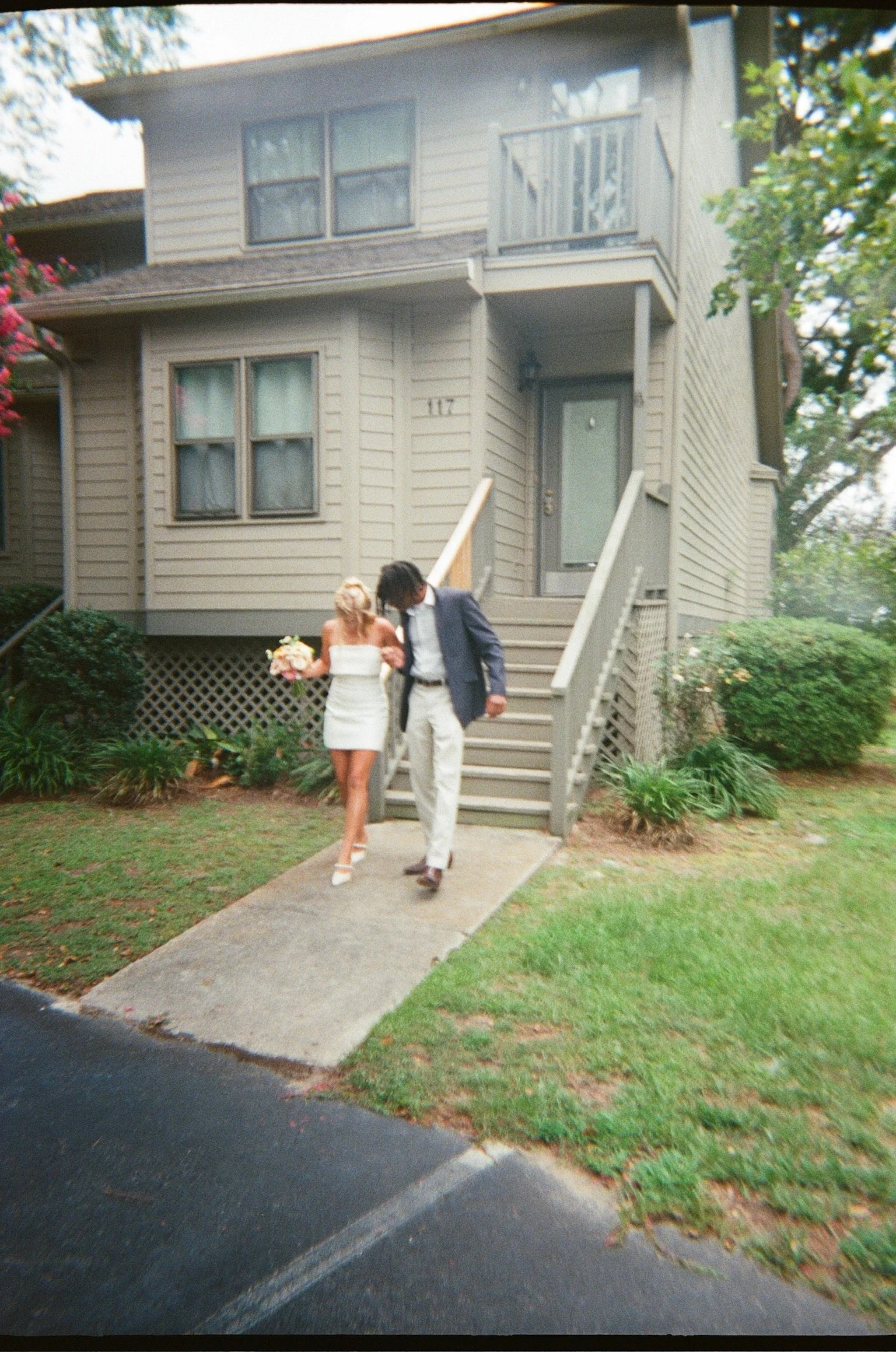 A bride in a white wedding dress holding a bouquet, walking down the stairs outside a beige house with a second-story balcony, assisted by a man in a dark suit.