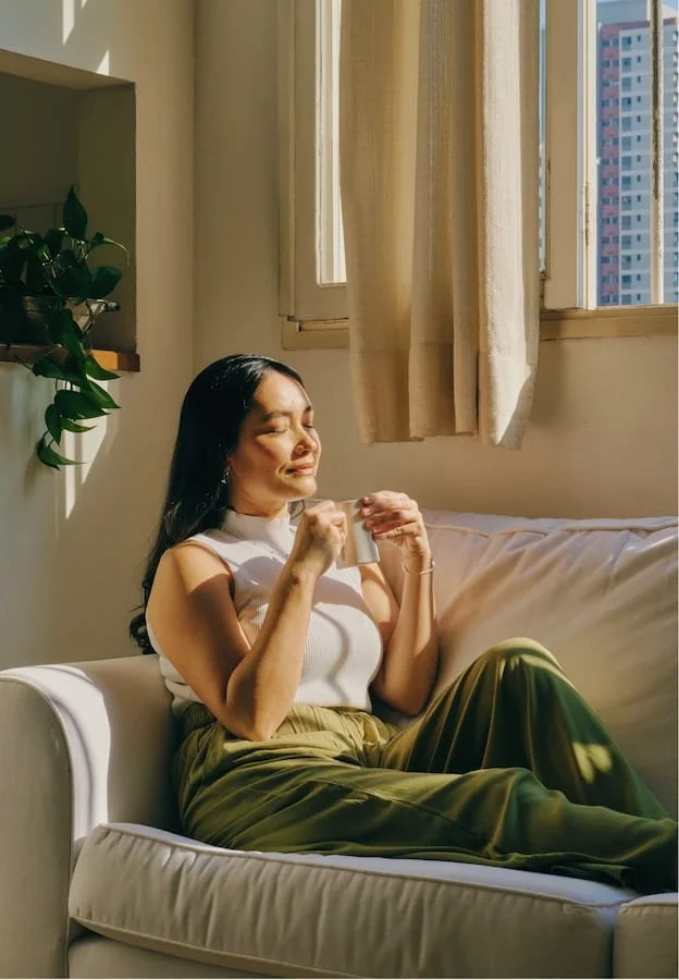 Woman sitting on a sofa beside a sunlit window, holding a mug with her eyes closed.