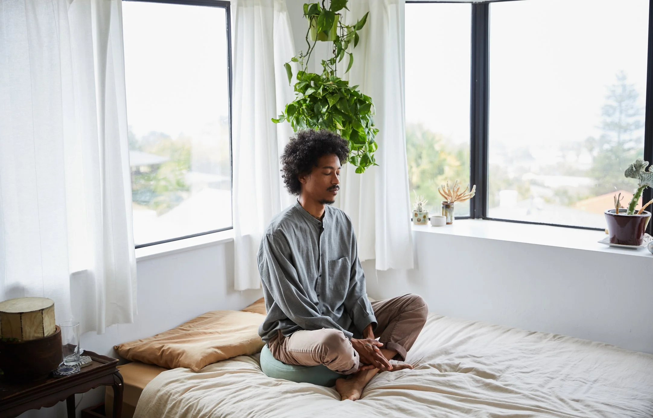 Man sitting cross-legged on a bed with his eyes closed, surrounded by natural light and hanging plants.